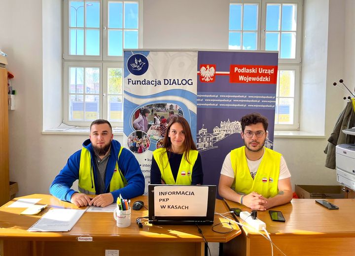 Trzy osoby w żółtych kamizelkach siedzą za stołem, za nimi są tablice Fundacji Dialog i Podlaskiego Urzędu Wojewódzkiego