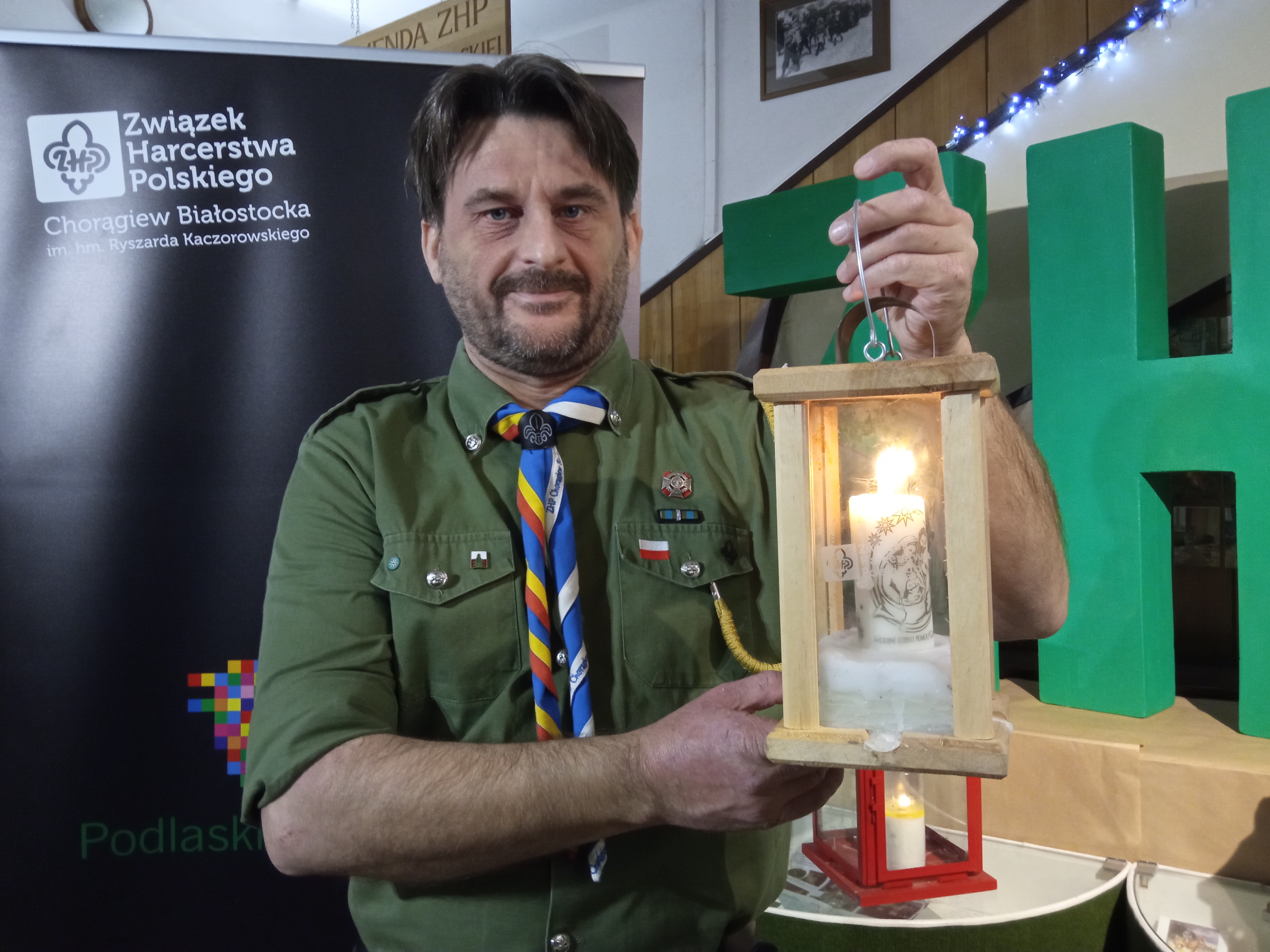 Mężczyzna w harcerskim mundurze trzyma lampion z zapalonym zniczem w środku