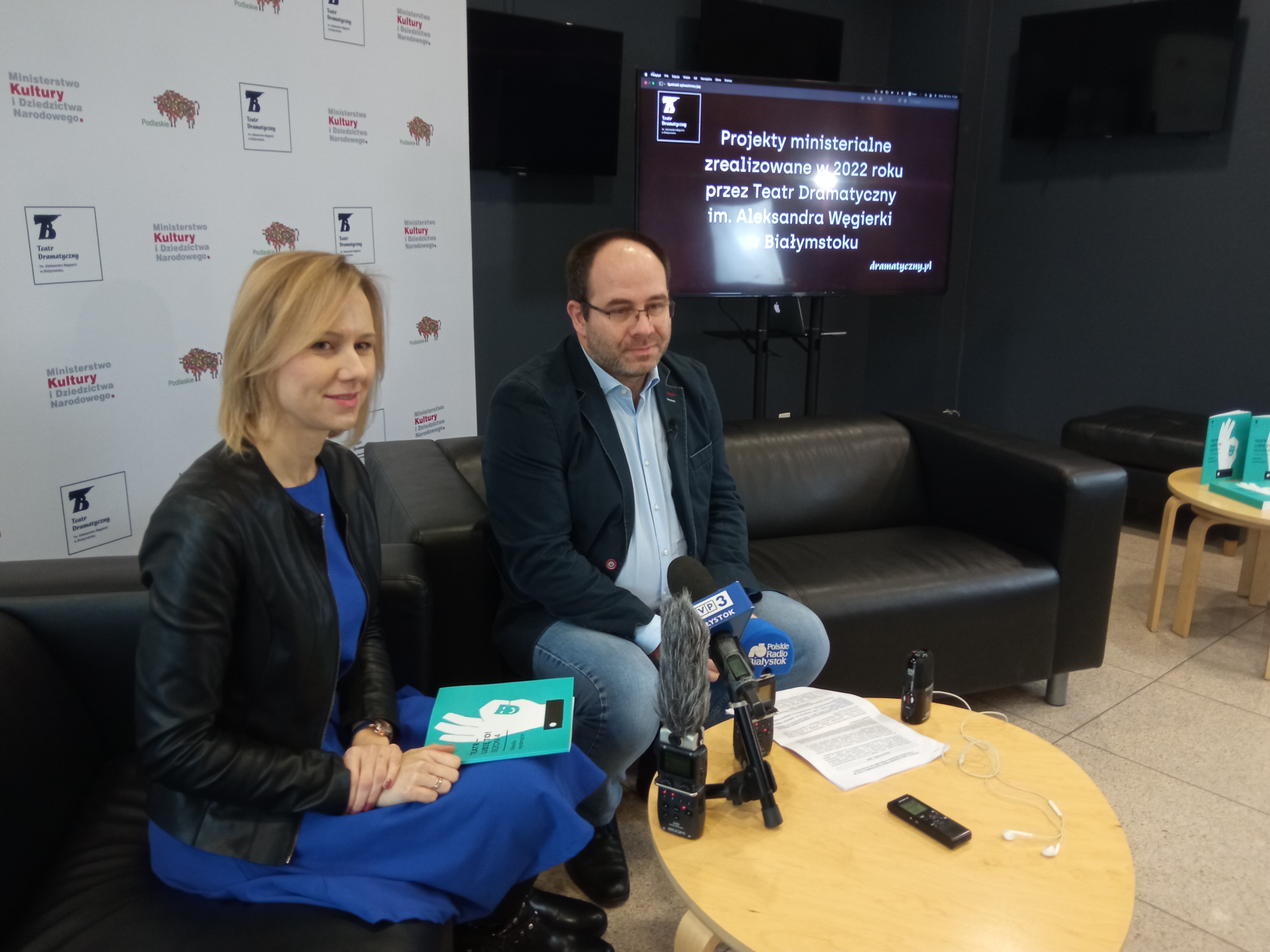 Kobieta i mężczyna siedzą na kanapie podzcas konferencji; na stole stoją mikrofony