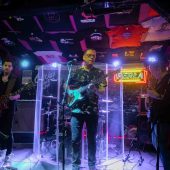 Zespół występuje na scenie, trzech mężczyzn gra na gitarze