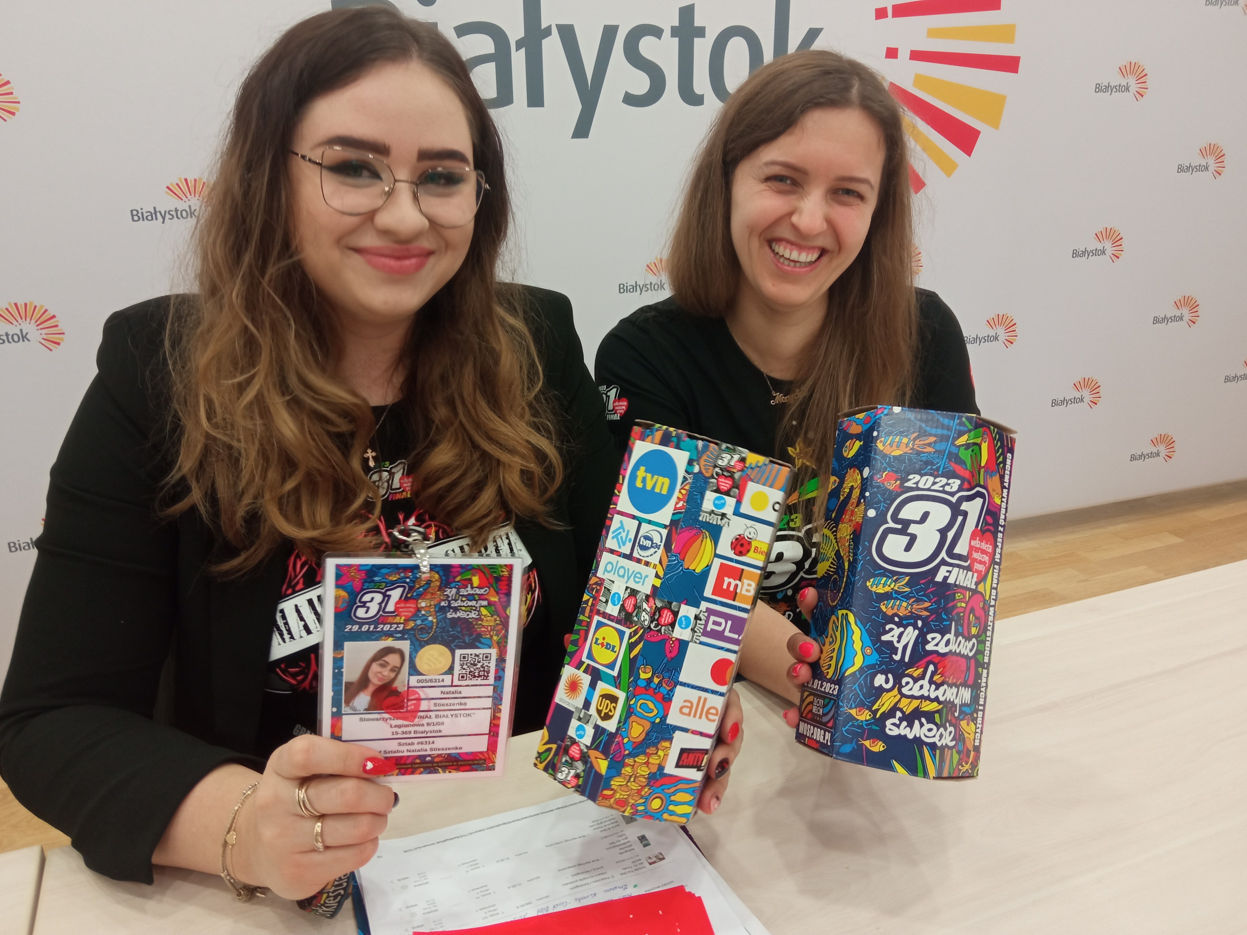 Dwie kobiety siedzą za stołem, trzymają puszki WOŚP i identyfikator