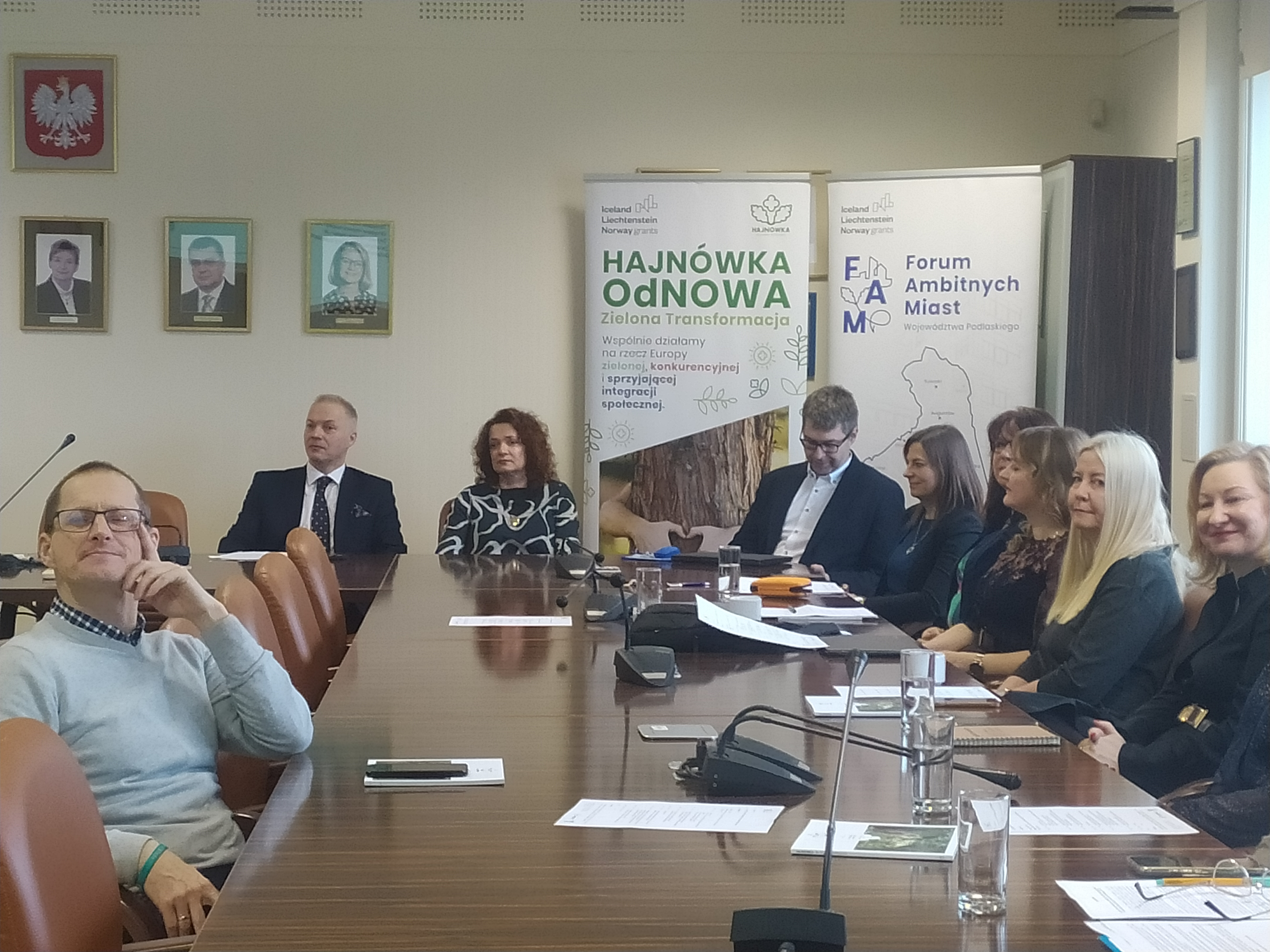 Elegancko ubrane osoby siedzą przy dużym stole, na którym leżą kartki