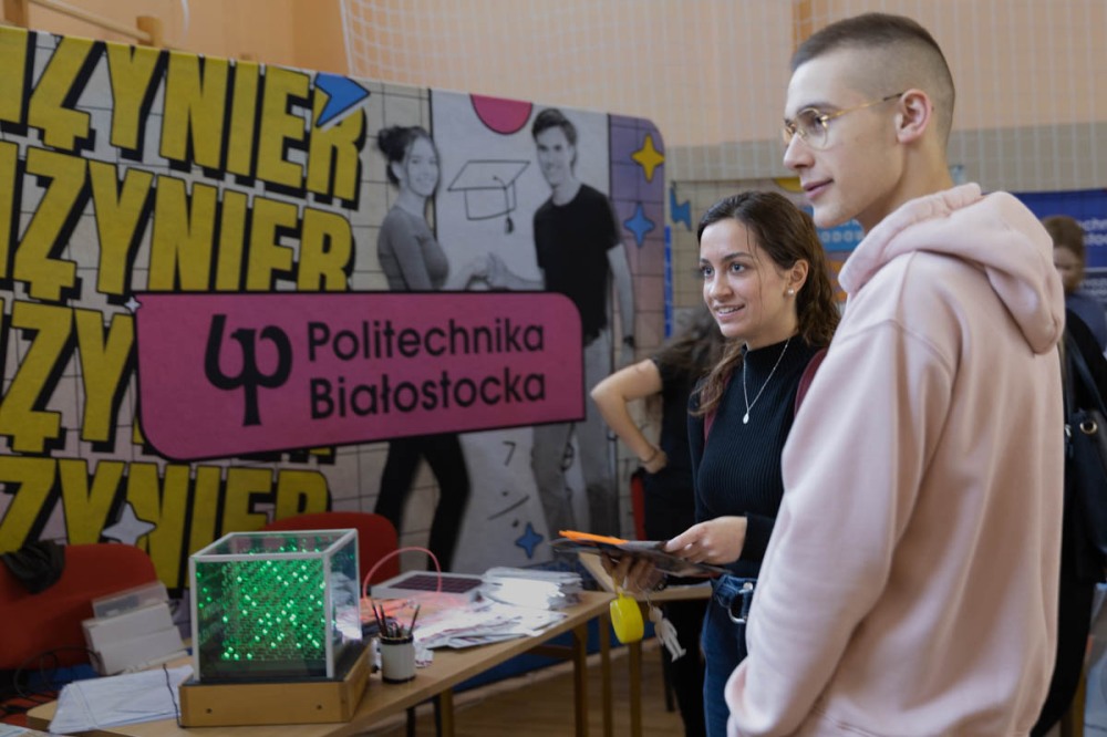 Dwoje młodych ludzi stoi przed stołem z maszyną i banerem Politechniki Białostockiej