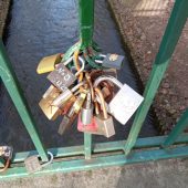 Kolorowe kłódki wiszą na balustradzie mostka; poniżej płynie rzeka