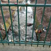 Kolorowe kłódki wiszą na balustradzie mostka; poniżej płynie rzeka
