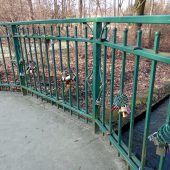 Kolorowe kłódki wiszą na balustradzie mostka; poniżej płynie rzeka
