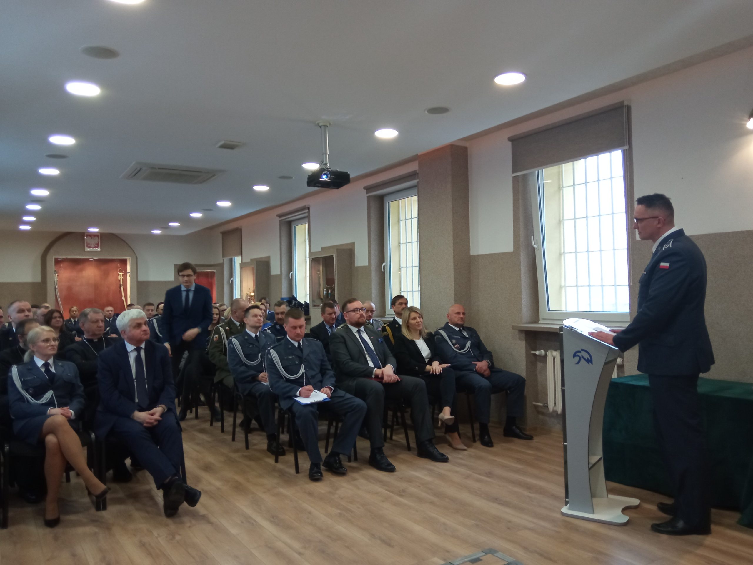 Funkcjonariusze w mundurach i osoby cywilne siedzą na auli i słuchają wykładu stojącego mężczyzny