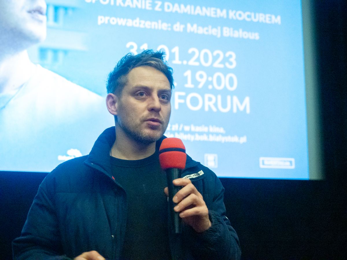 Młody mężczyzna stoi na tle wyświetlanego filmu i trzyma mikrofon