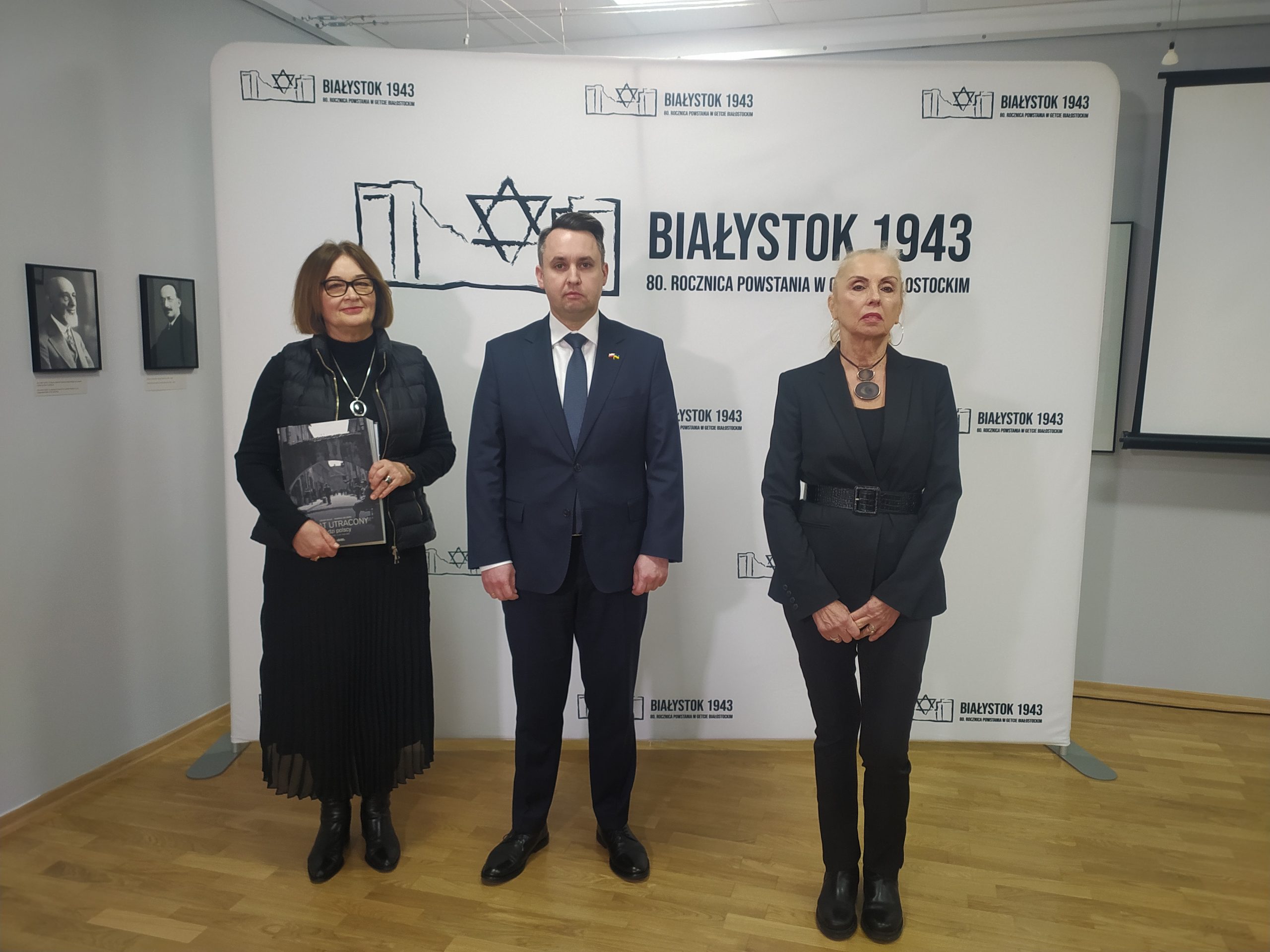 Dwie kobiet y i mężczyzna stoją przed tablicą z logiem, na którym jest frmagnet muru i obok napis Białystok 1943.