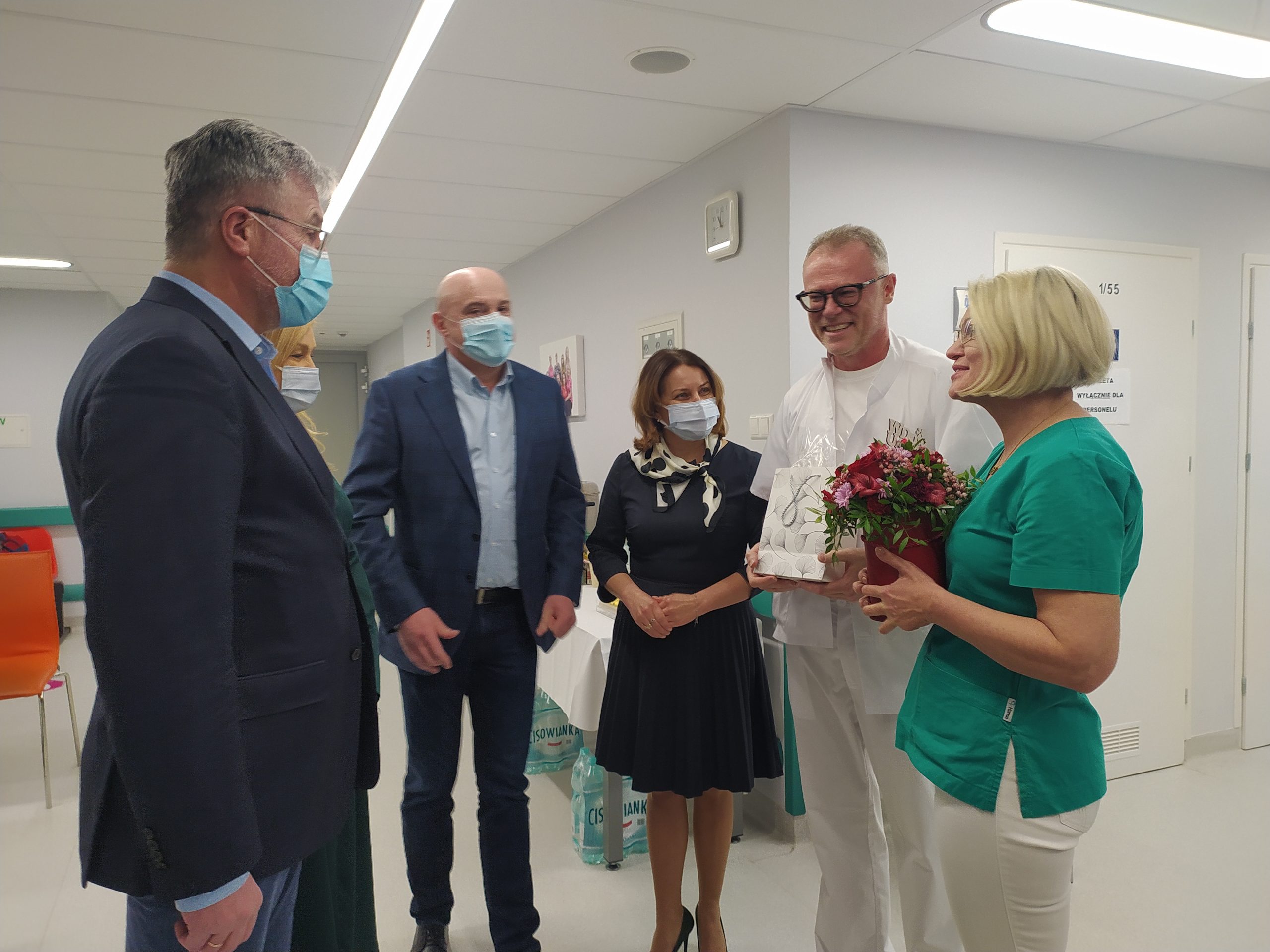Pielęgniarka i lekarz trzymają prezent i kwiaty, obok nich stoją trzy osoby elegancko ubrane