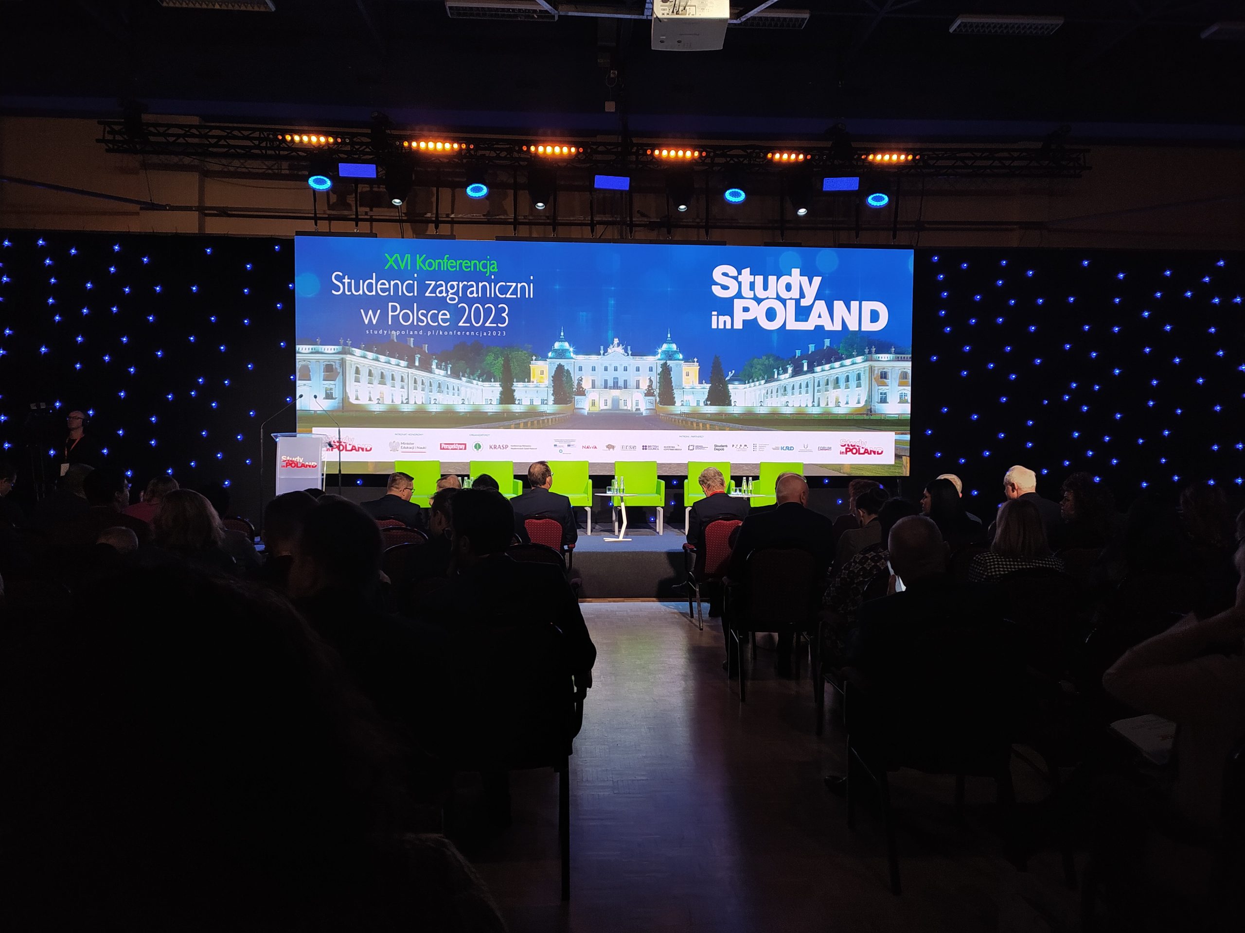 Ciemna sala, na której końcu jest duży telebim z wyświetlaną grafiką z naposem "Study in Poland"