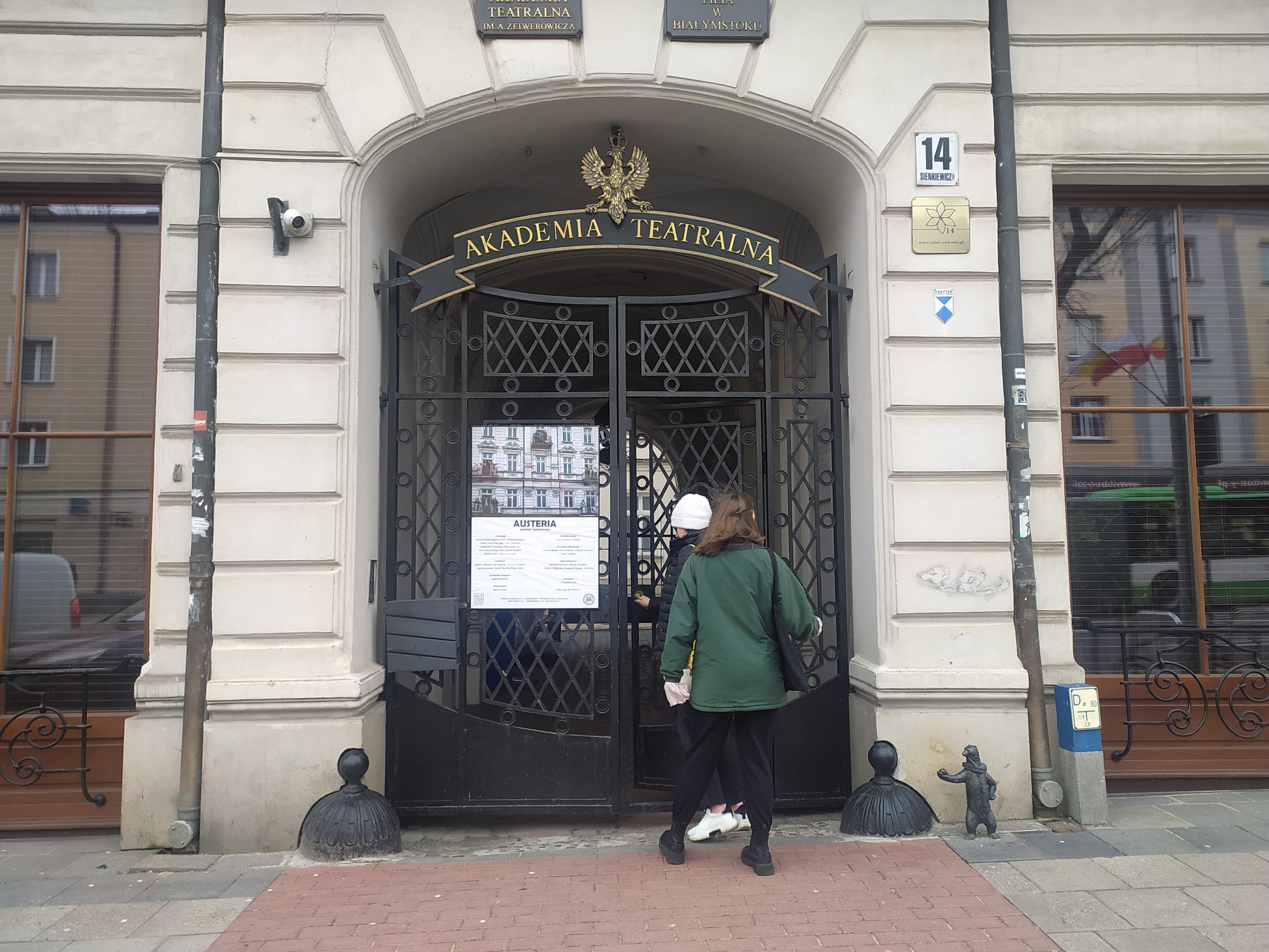 Dwie osoby wchodzą przez bramę, nad którą wisi napis Akademia Teatralna