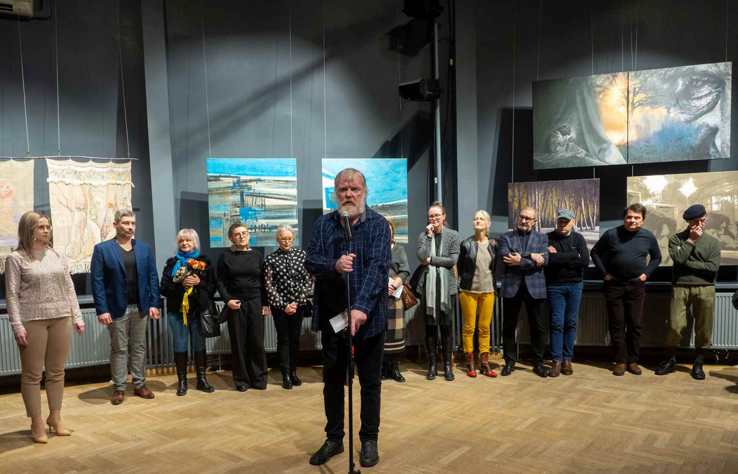 Grupa kilkunastu osób stoi pod ścianą, na której wiszą obrazy, na środku przed mikrofonem stoi mężczyzna.