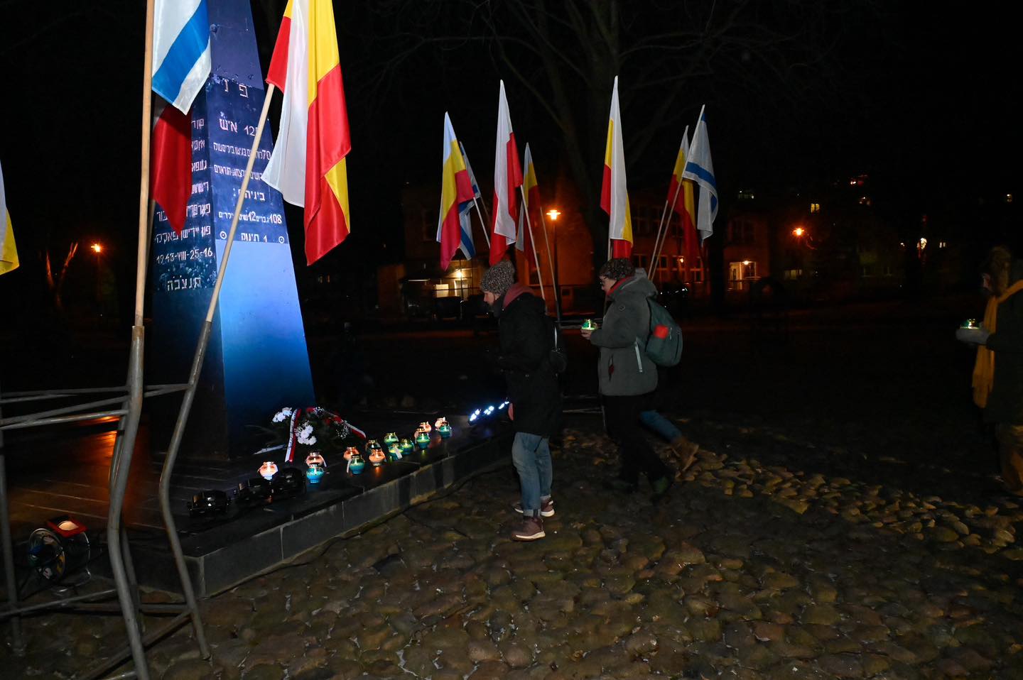 Dwóch ludzi podchodzi ze zniczami do pomnika, obok którego stoją flagi