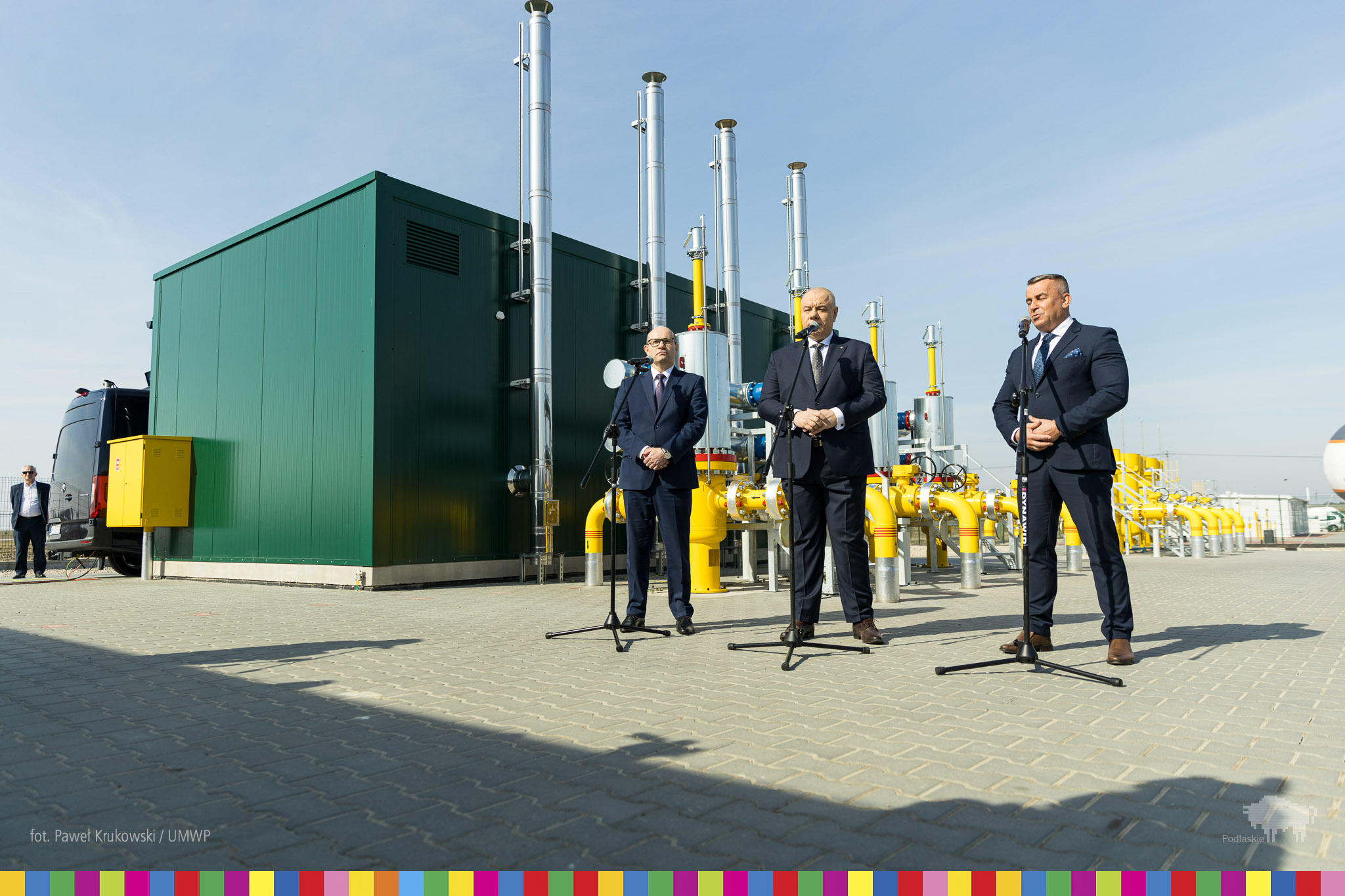 Trzech mężczyzn stoi przed mikrofonami na podwórku przed zieloną halą i żółtymi rurami.