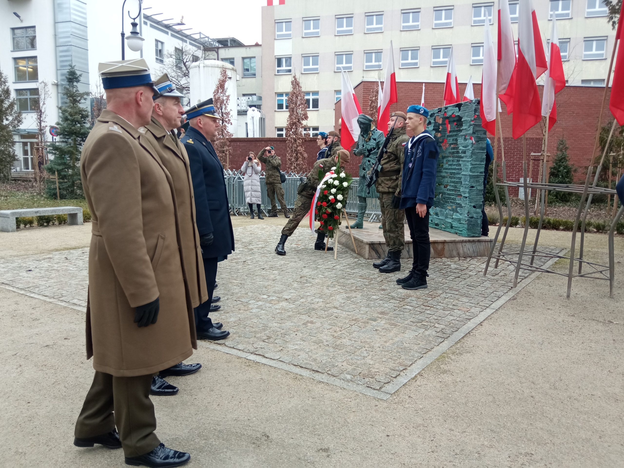 Żołnierz składa winiec pod pomnikiem, przy pomnu stoi warta, przed pomnikiem w szeregu stoją przedstawiciele władz