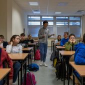 Uczniowie siedzą przy stołach słuchają wykładu, pomiędzy stołami idzie wykładowca