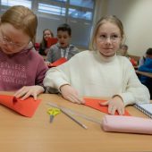Dziewczynki siedzą przy stole w klasie skłądają kolorowy papier, w tle przy stołach siedzą inne dzieci