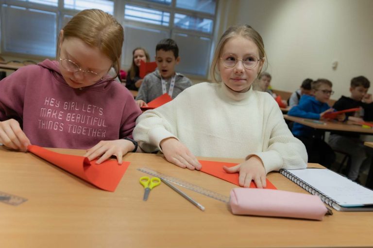 Dziewczynki siedzą przy stole w klasie skłądają kolorowy papier, w tle przy stołach siedzą inne dzieci