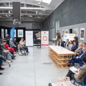 Duża hala, w której po lewej stronie siedzą ludzie na krzesłach, a na przeciw nich za stołem siedzą uczestnicy konferencji.