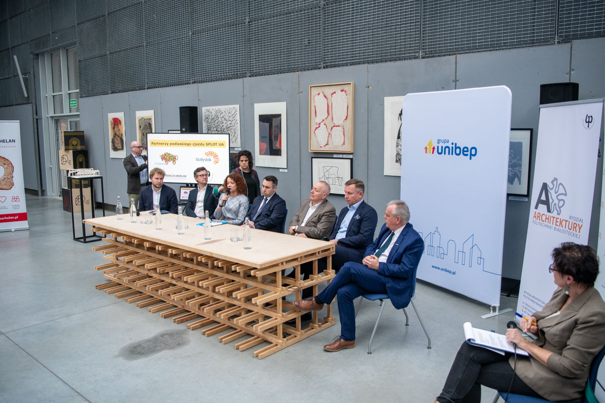 W dużej hali przy stole siedzą ludzie w trakcie konferencji.