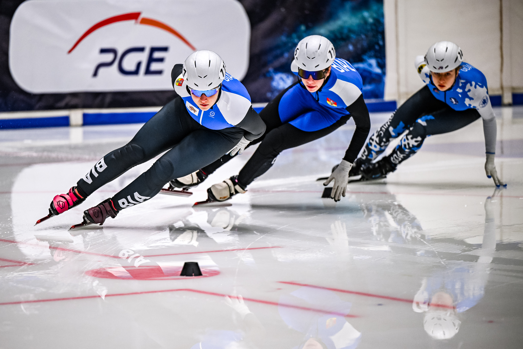 Trzy osoby w sportowych strojach i kaskach jadą na łyżwach.