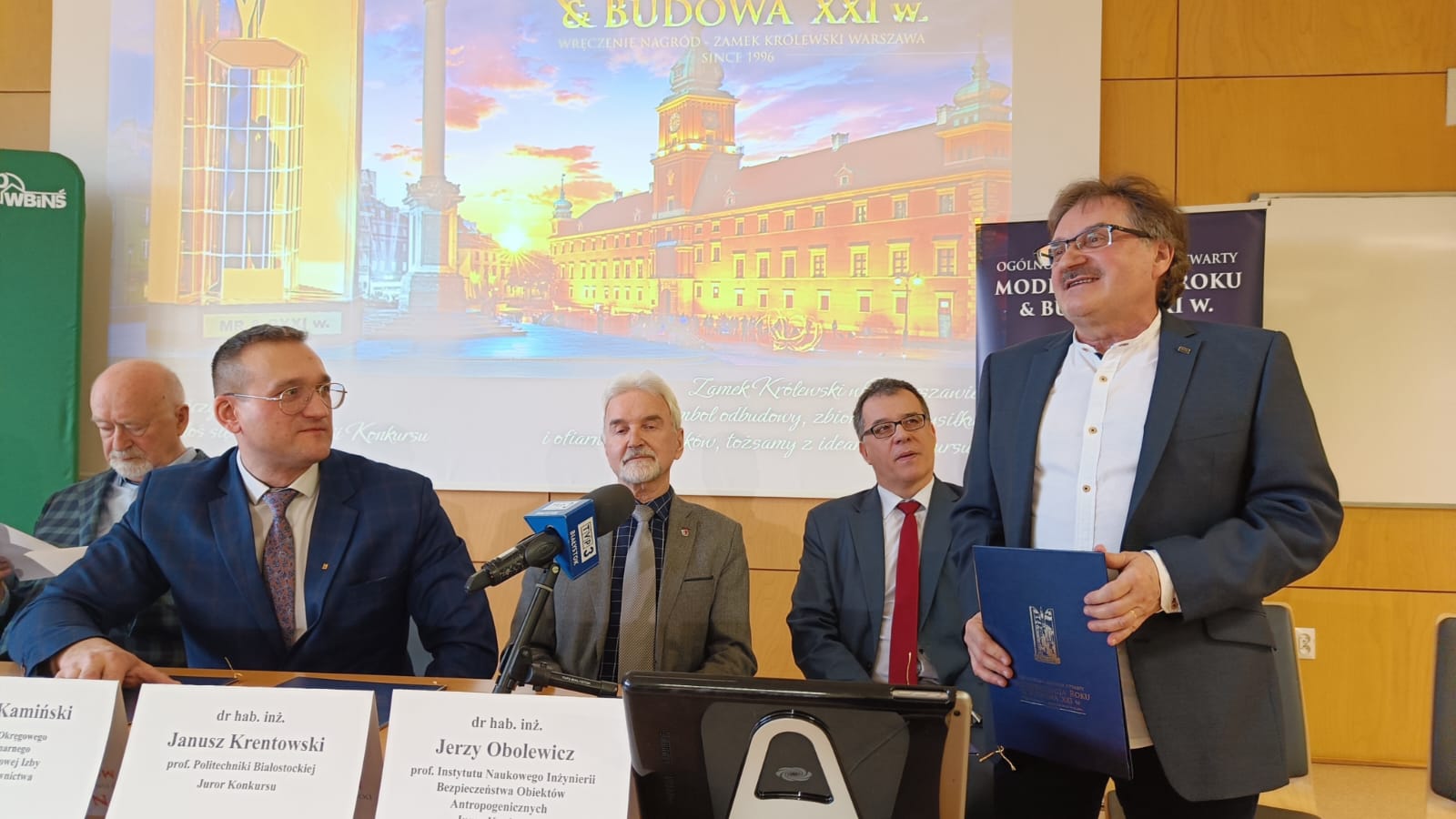 Czterech mężczyzn siedzi za stołem, za nimi wyświetlana jest prezentacja na rzutniku. Jeden mężczyzna stoi i przemawia.
