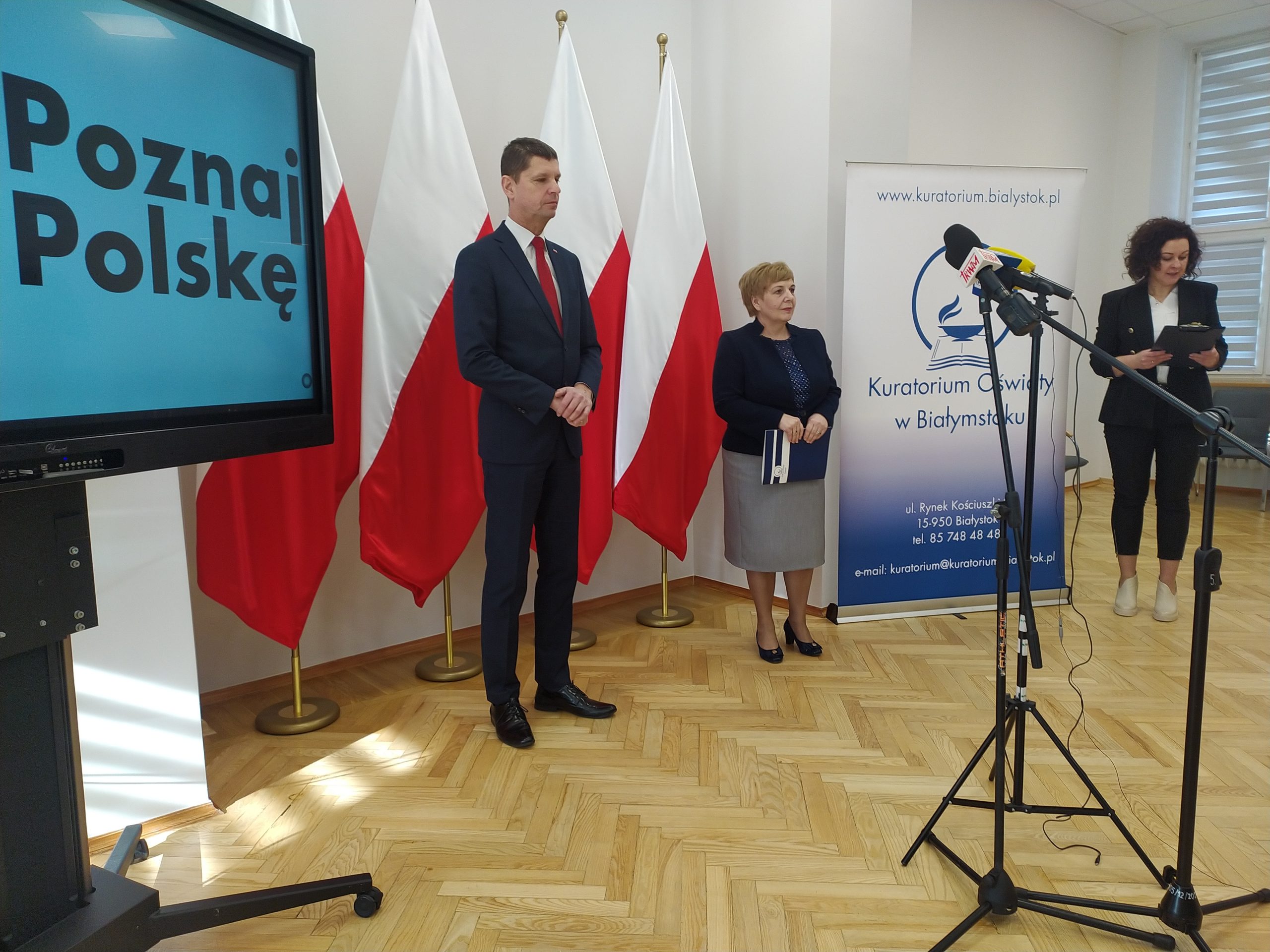 Dwie osoby stoją przed mikrofonami na tle flag Polski.