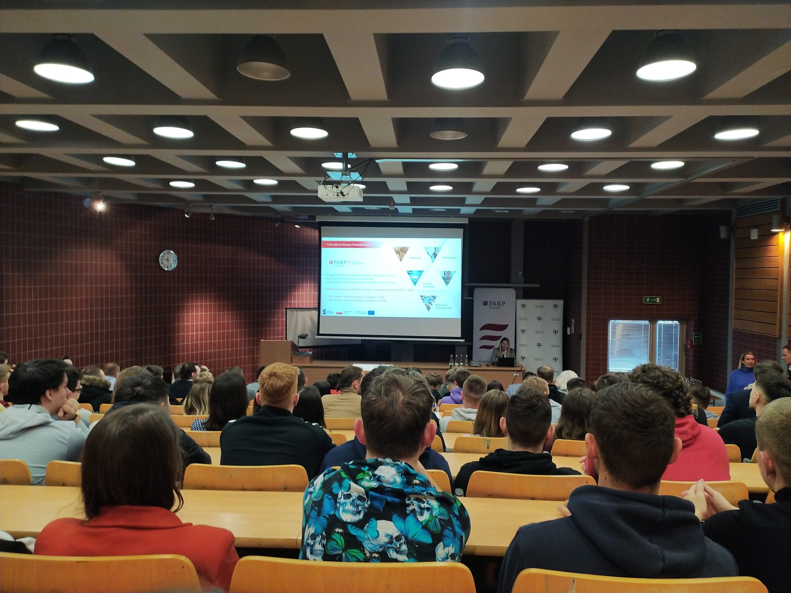 Młodzi ludzie siedzą w auli przed telebimem.