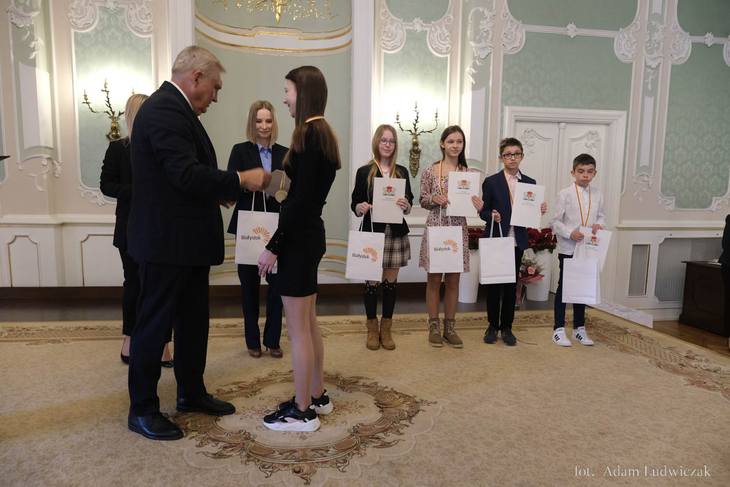 Mężczyzna podaje rękę młodej dziewczynie, z tyłu stoi młodzież z dyplomami.