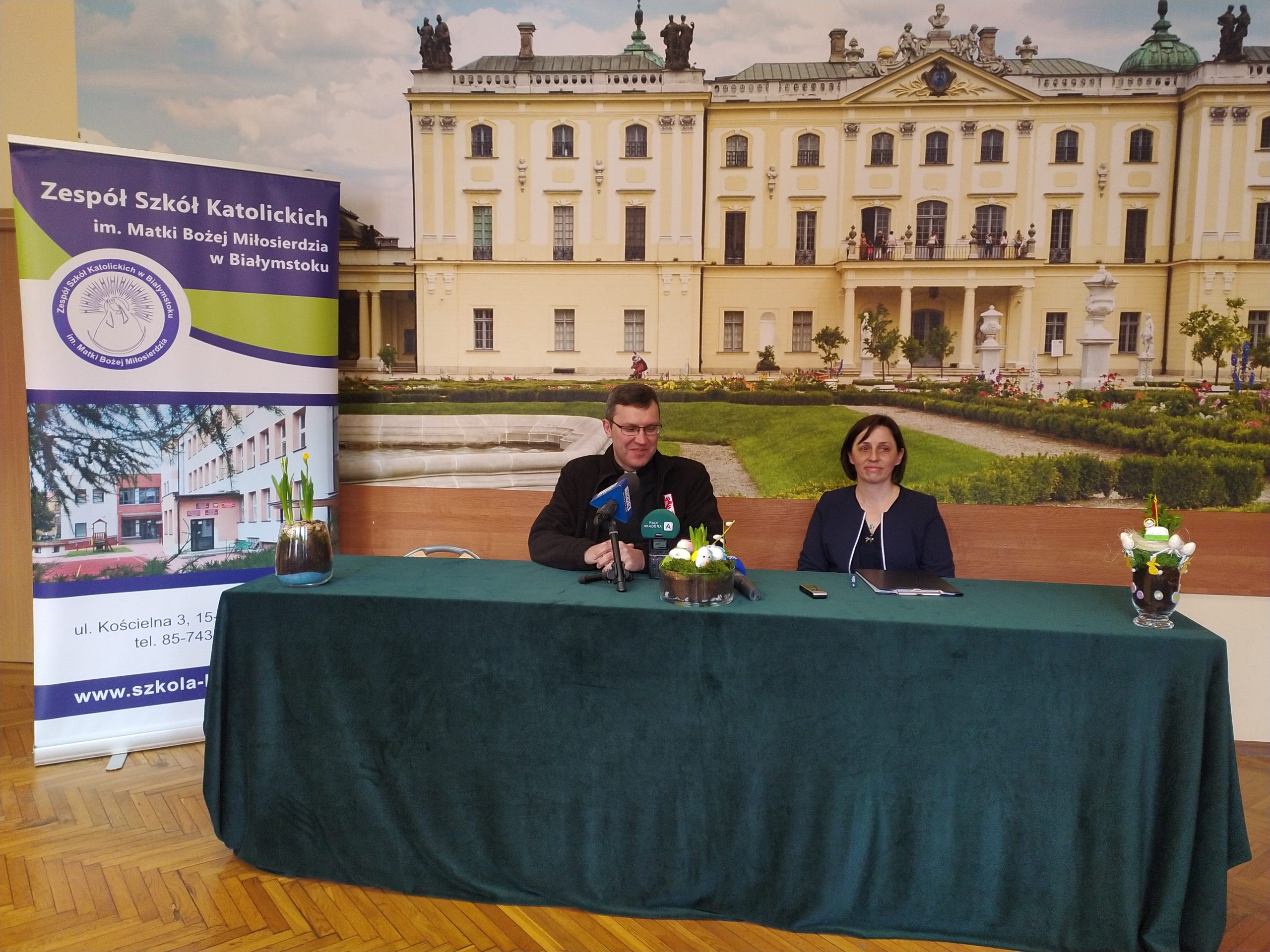Ksiądz i kobieta siedzą za stołem przed mikrofonami.