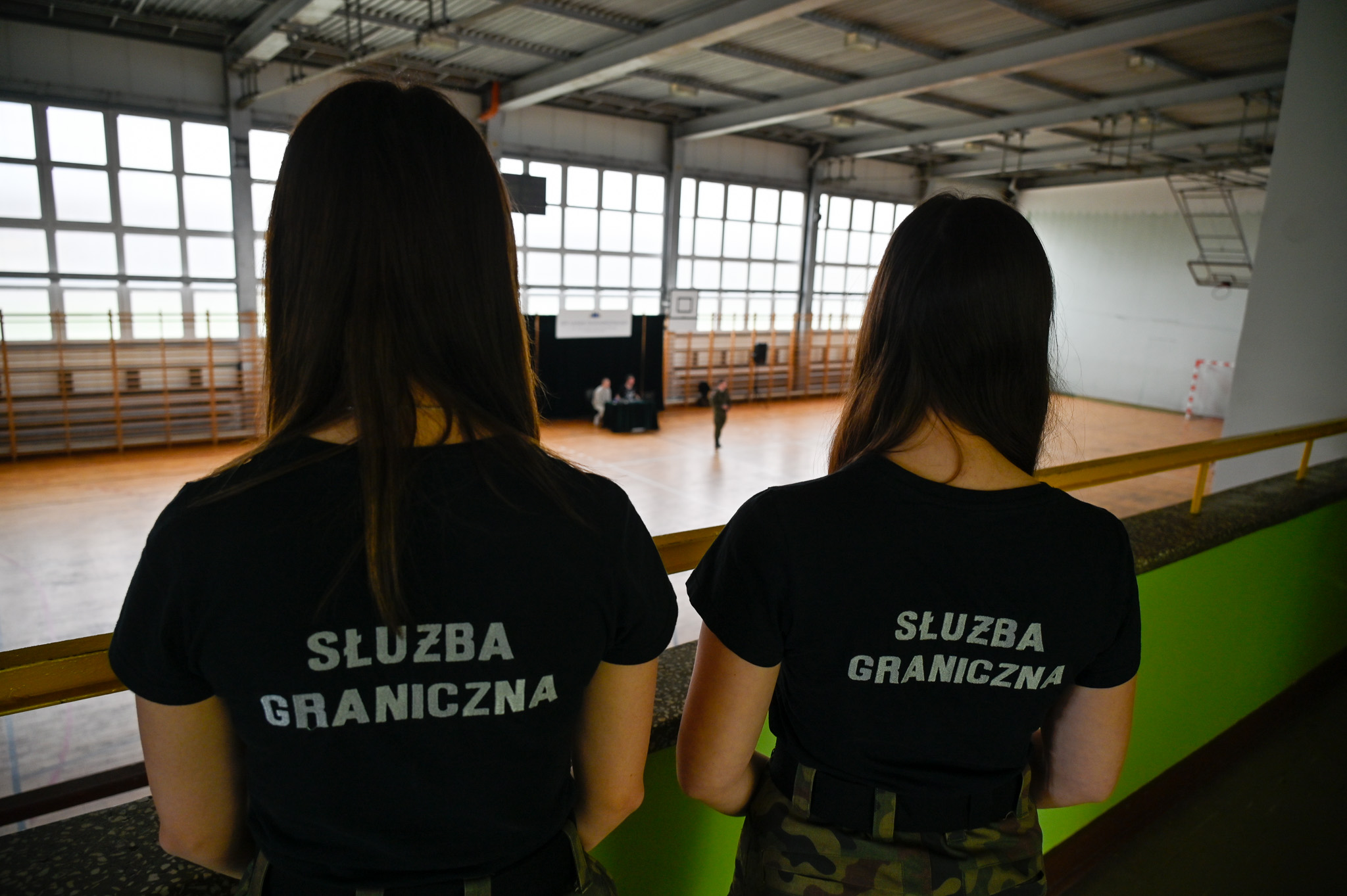 Dwie dziewczyny stoją tyłem, na koszulkach mają napis: służba graniczna. W tle widać szkolną salę sportową.