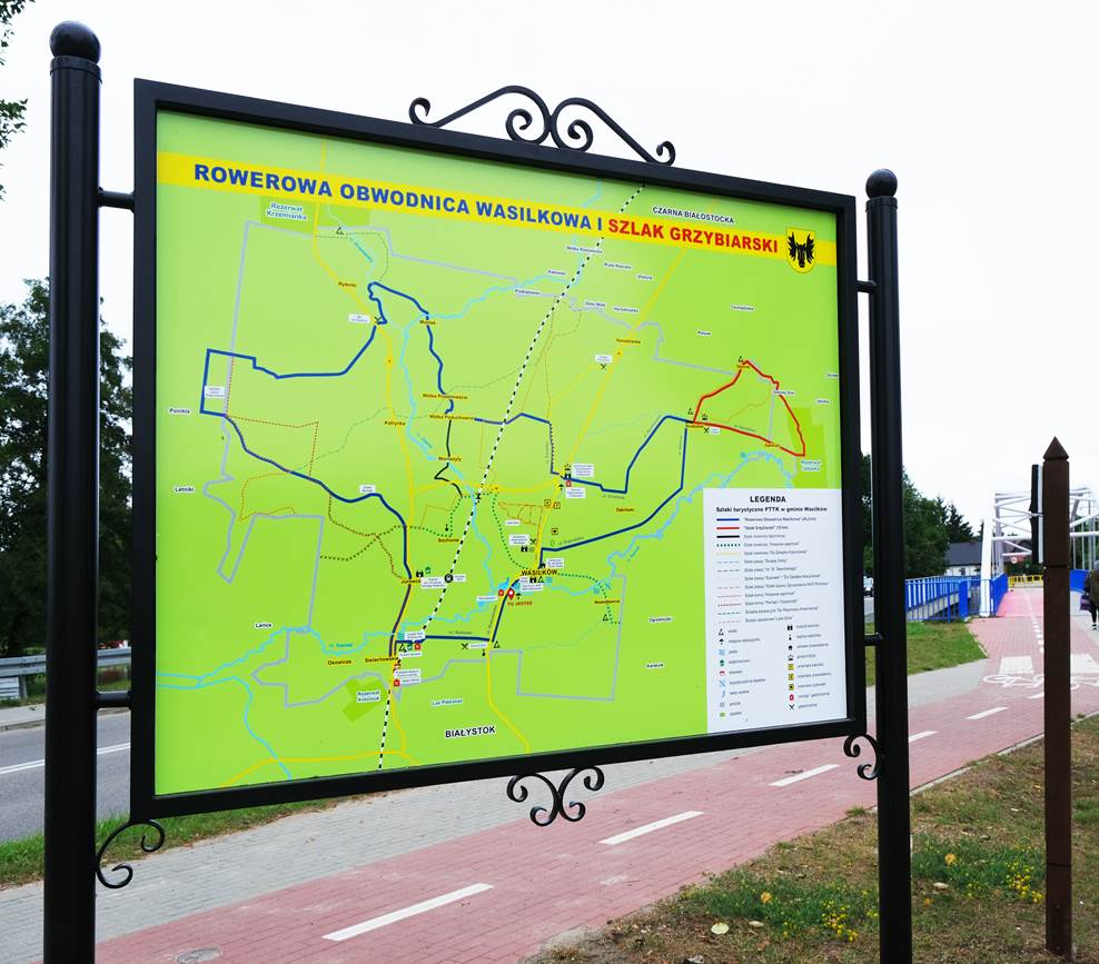 Mapa na zielonym tle umieszczona na stelażu obok ścieżki rowerowej.