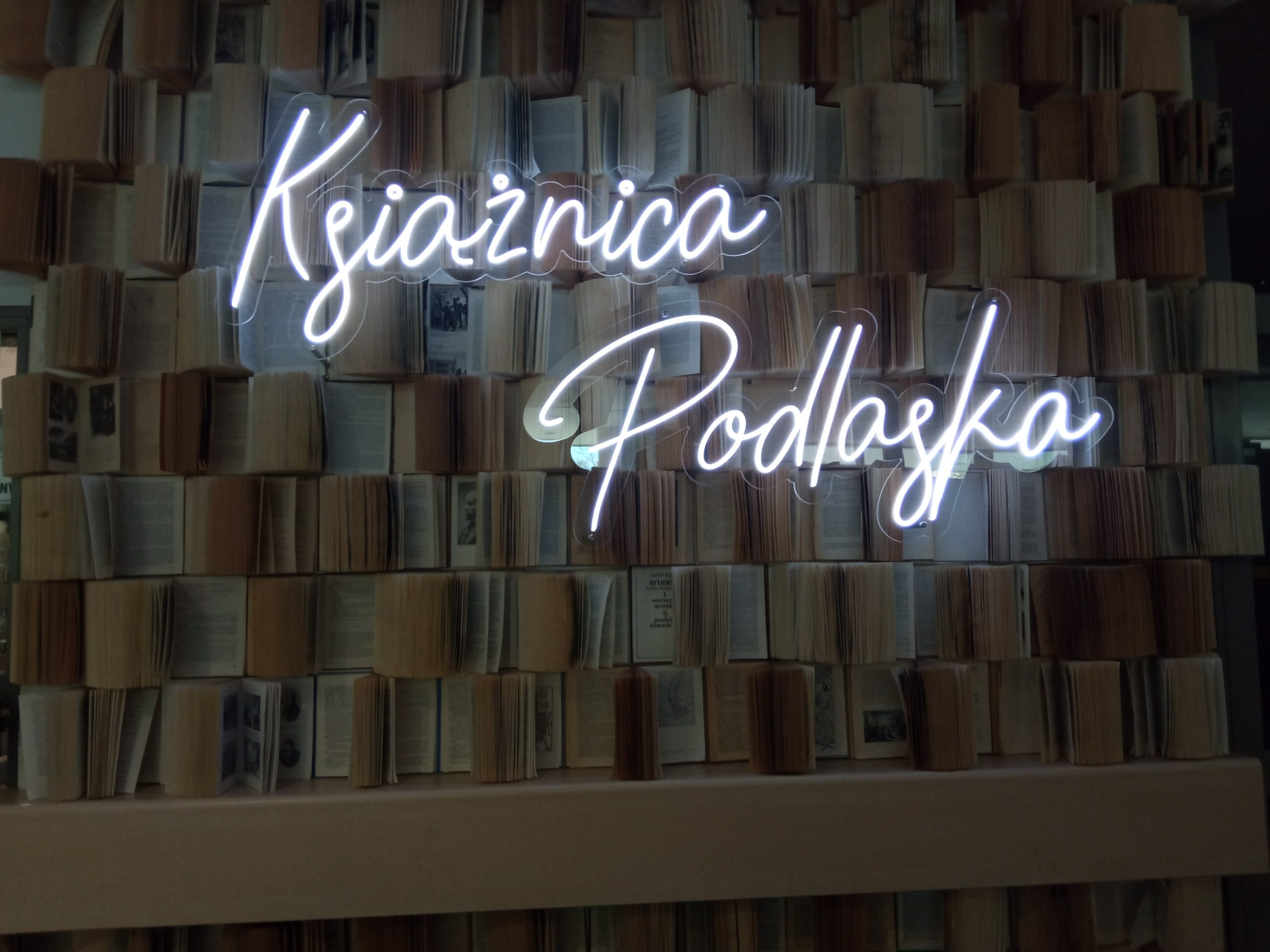 Napis neon Książnica Podlaska na ścianie, na tle książek