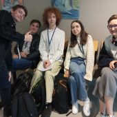 Grupa uczniów wraz z nauczycielką siedza n krzesłach na korytarzu