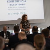 Kobieta mówi do mikrofonu, obok siedzą inne osoby, przed nimi siedzi kilkadziesiąt ośób