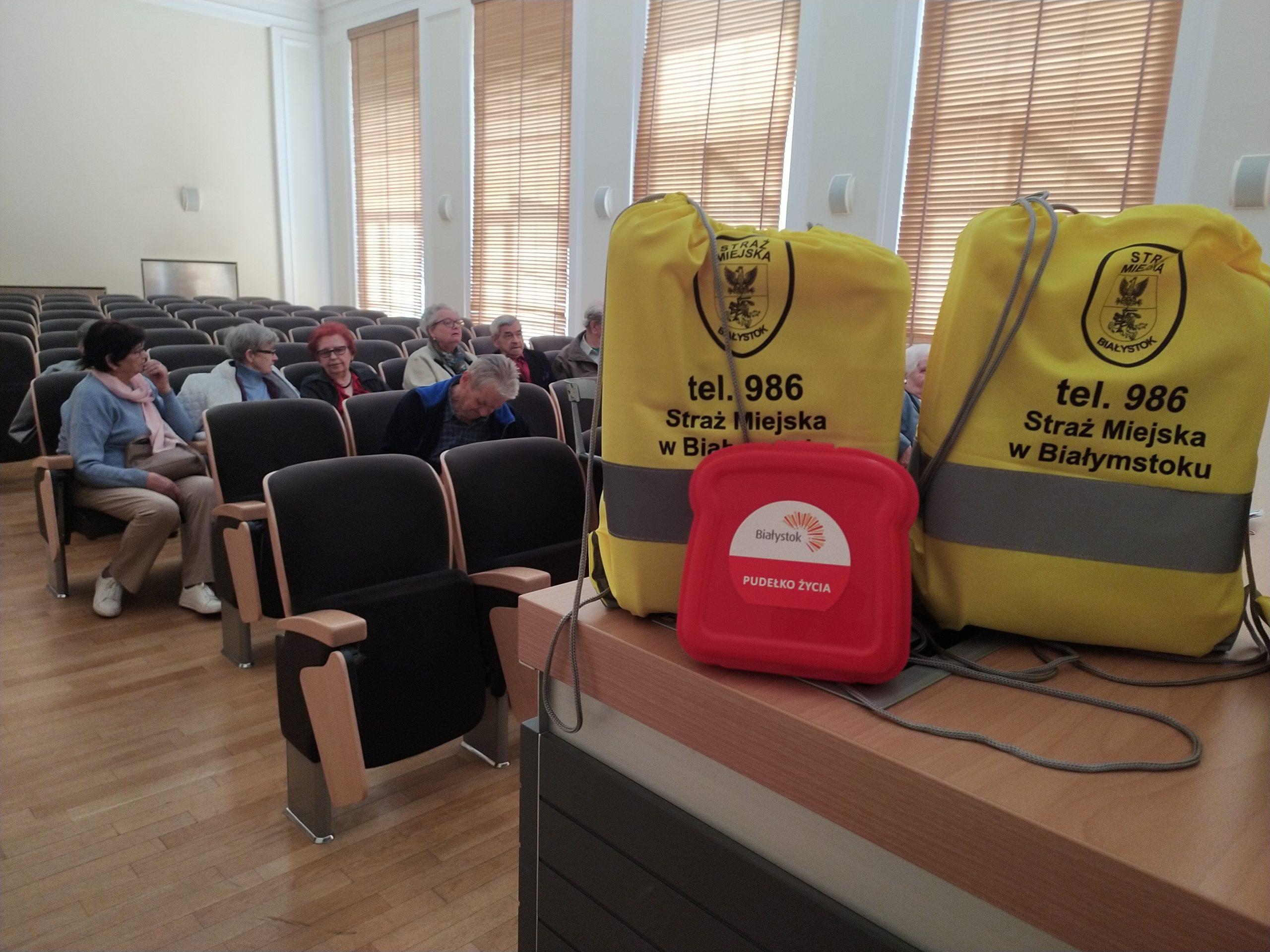 Na pierwszym tle torby z napisem straż miejska i pudełko życia, a w tle seniorzy siedzą na sali konferencyjnej.