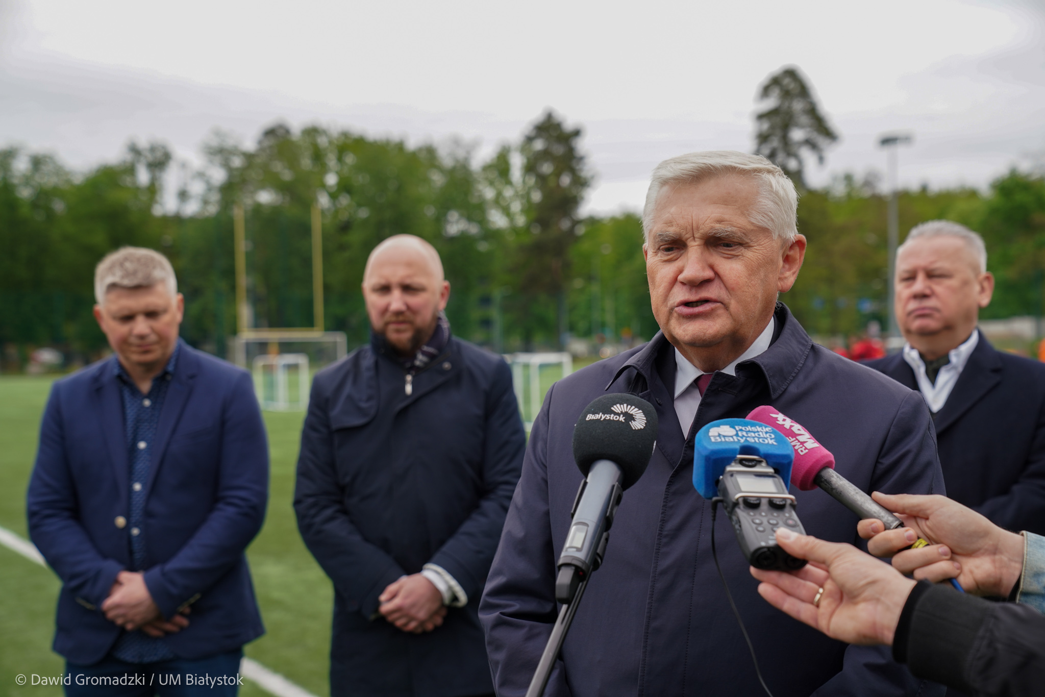 Mężczyzna stoi na pierwszym planie przed mikrofonem; Z tyłu stoi trzech męźczyzna