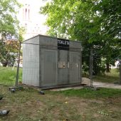 Szary budynke ogrodzony siatką, odsłonięte fundamenty, w tle drzewa