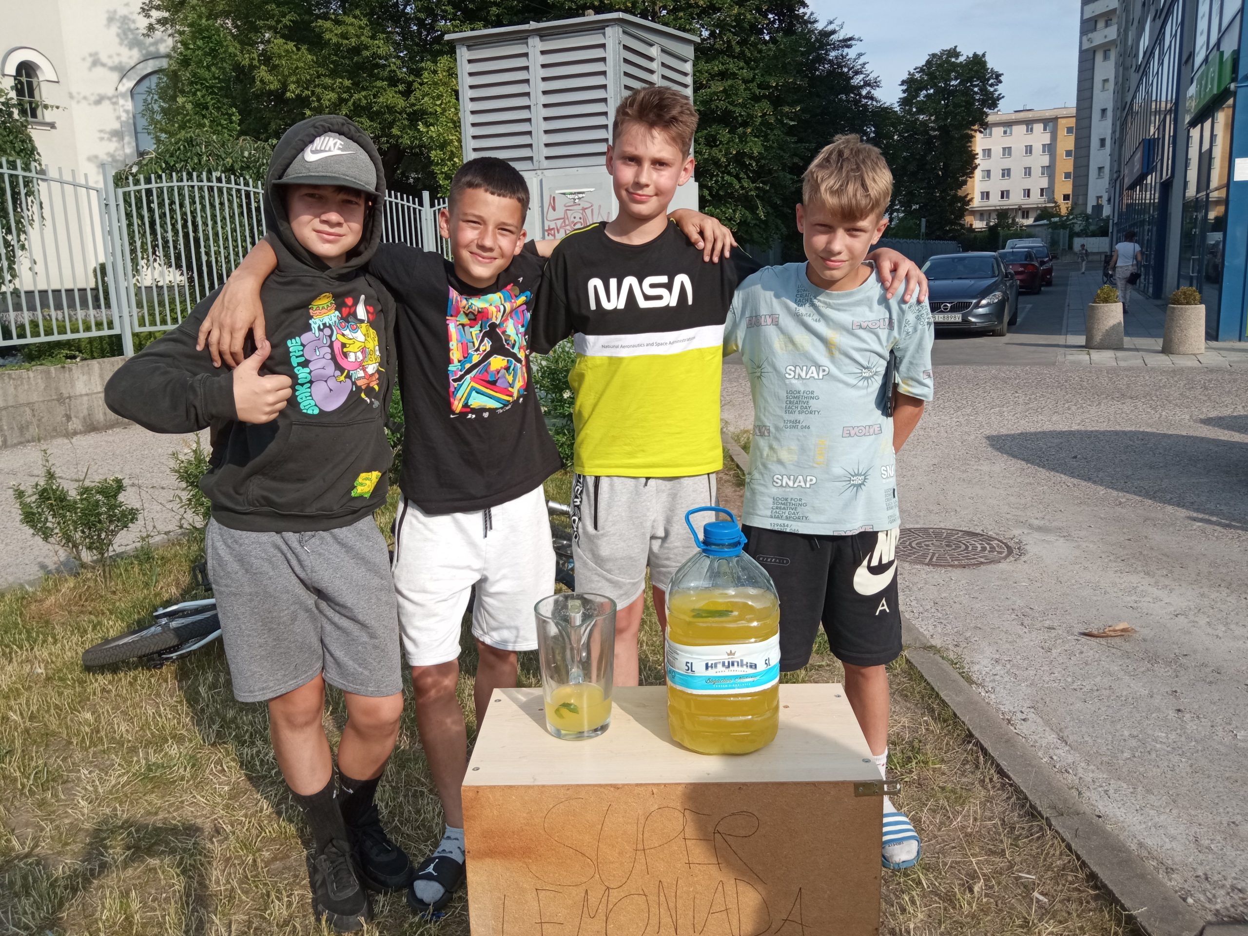Na stoliku st oi duża butelka z lemoniadą, obok dzbanek, za stolikiem stoi czterech młodych chłapaków, uczniowie