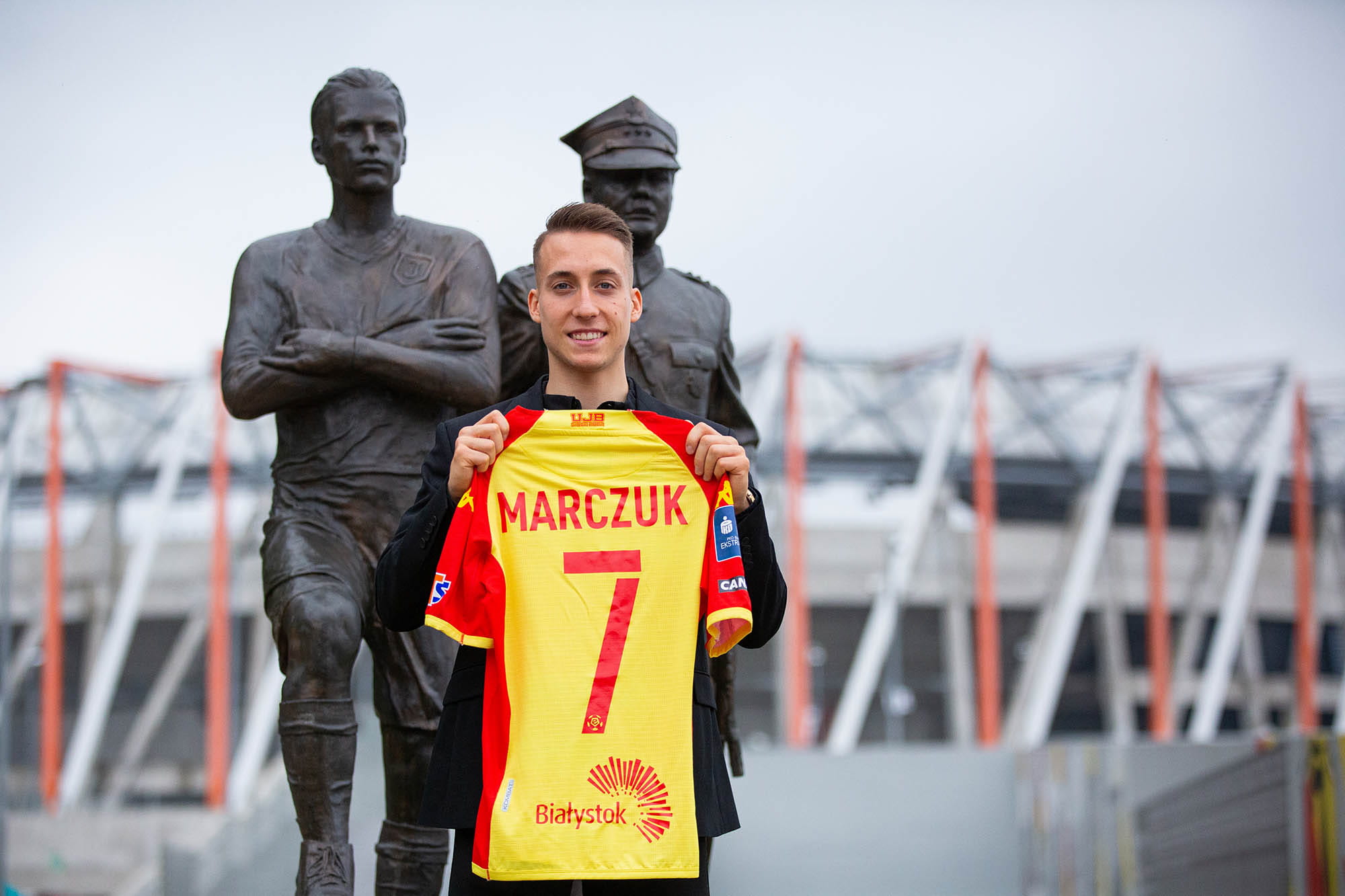 Młody mężczyzna trzyma w rękach koszulkę klubu Jagiellonia Białystok, stoi przed pomnikiem obok stadionu