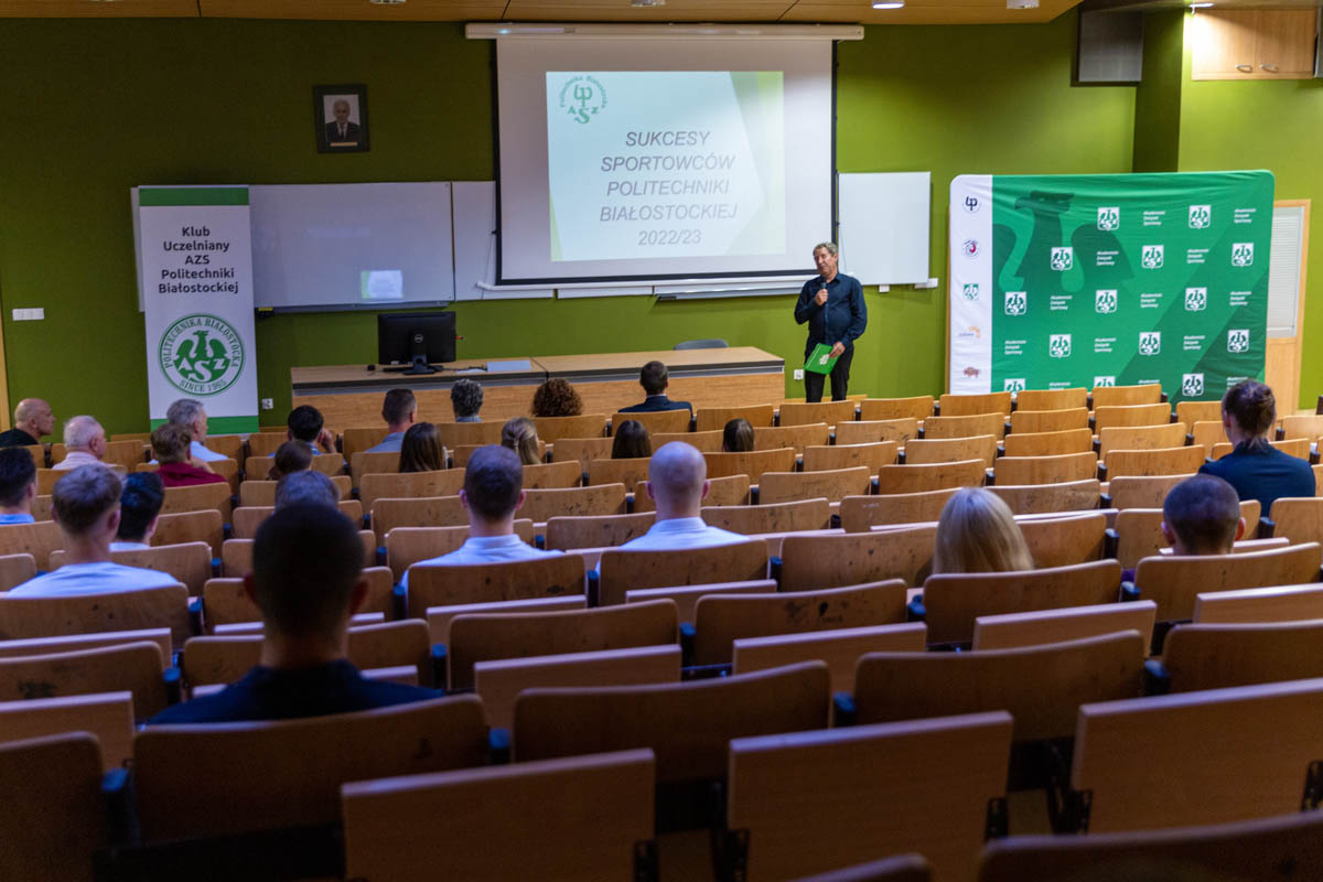 Na auli siedzą ludzie, z mównicy przemawia mężczyzna.