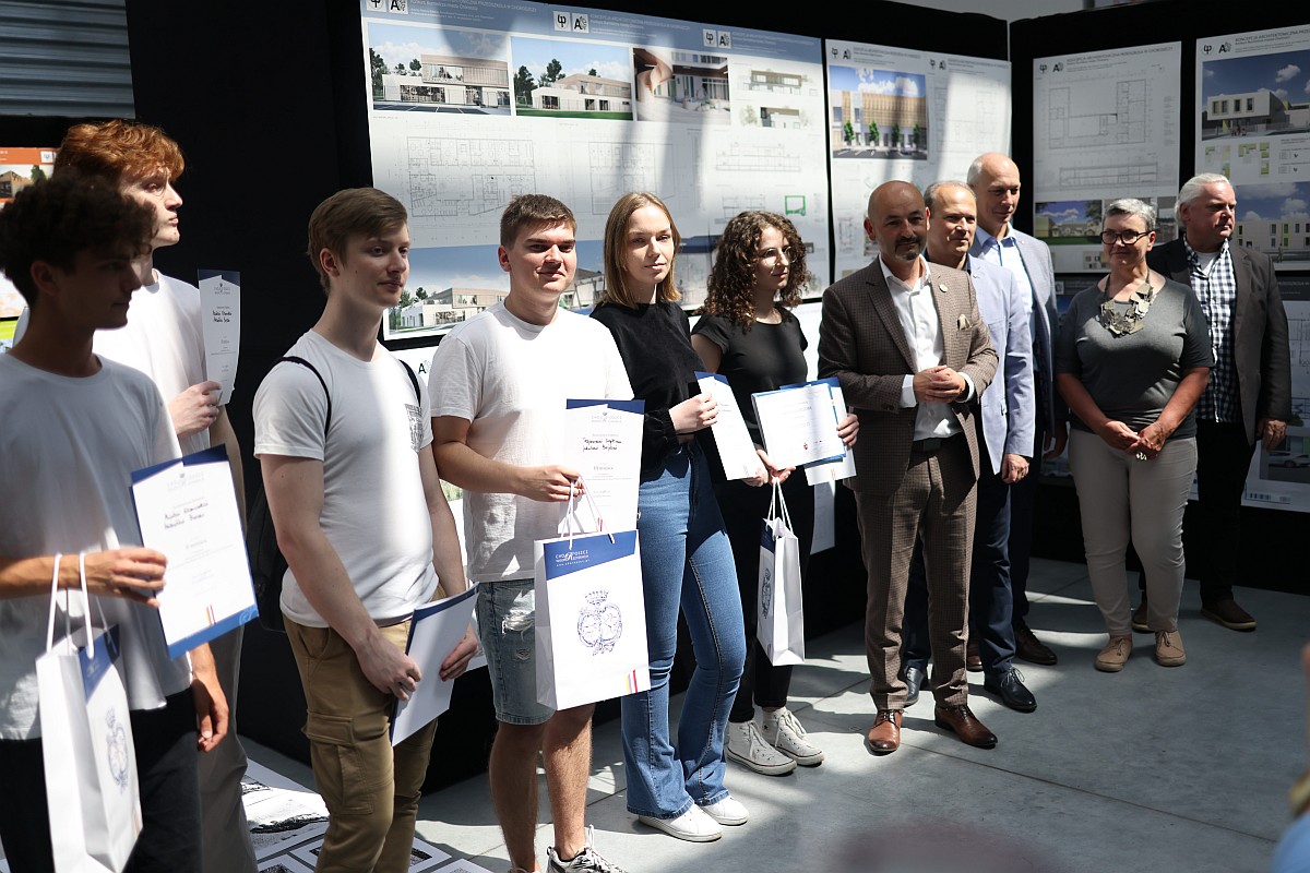 Ludzie pozują do zdjęcia na tle projektów architektonicznych wiszących na ścianie.