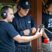 Policjant strzela na strzelnicy, a drugi mężczyzna pokazuje my telefon.