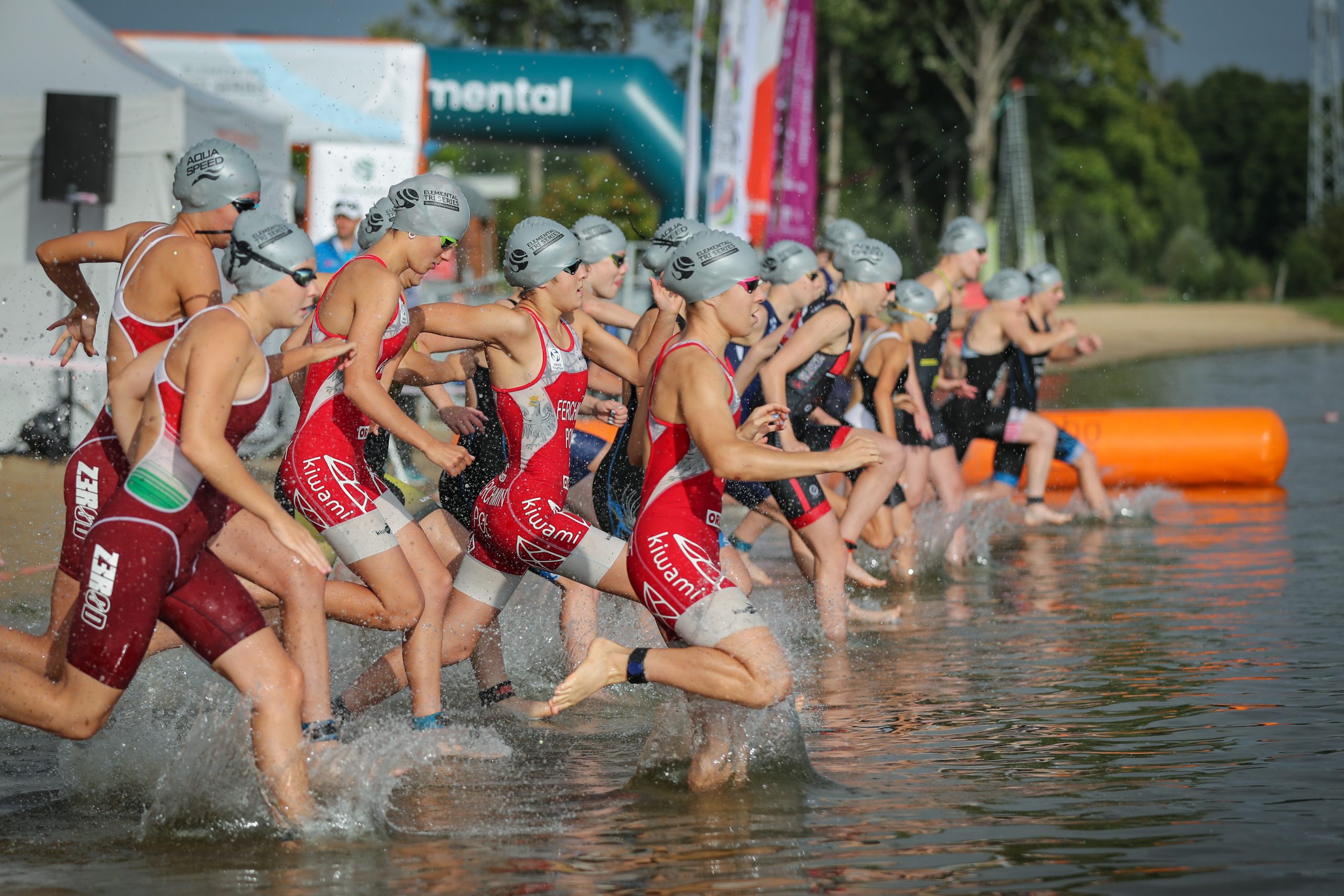 Grupa kobiet w sportowych strojach oraz czepkach i okularach pływackich wbiegają do wody