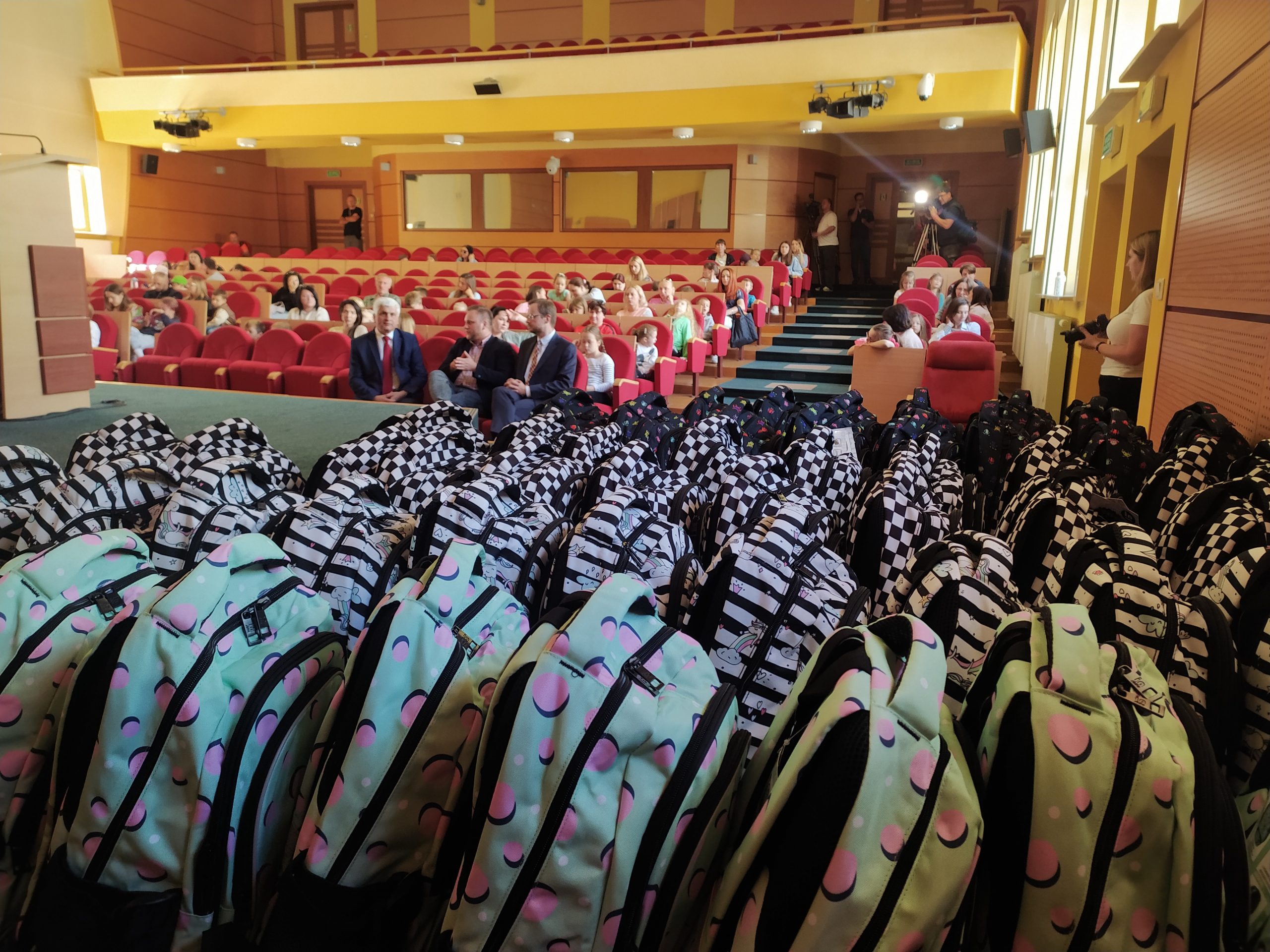 Na pierwszym planie plecaki szkolne, w głębi sala z konferencyjna, w której siedzą ludzie.