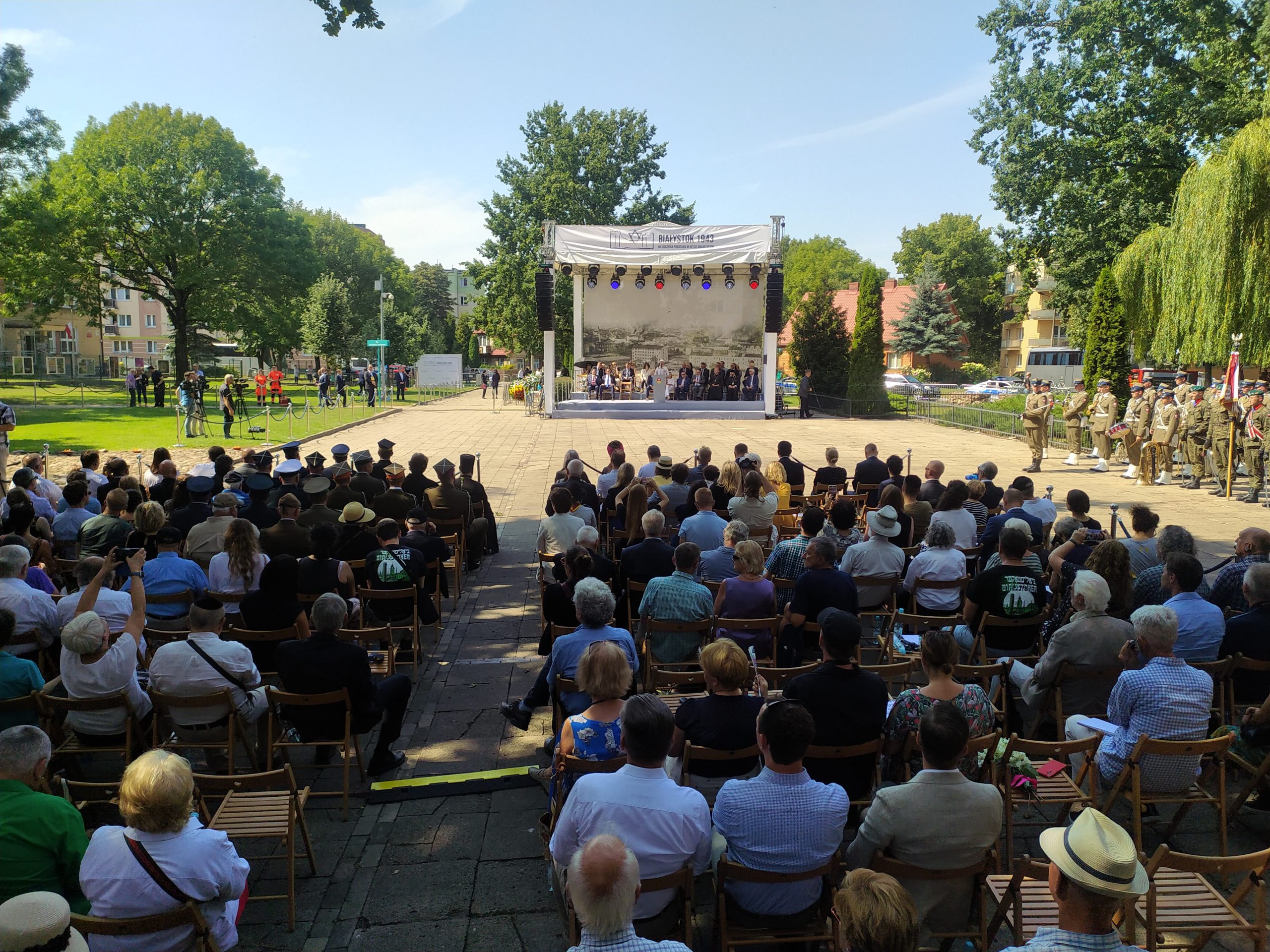 W tle widać scenę, a na pierwszym planie ludzie siedzą na krzesłach przed sceną.