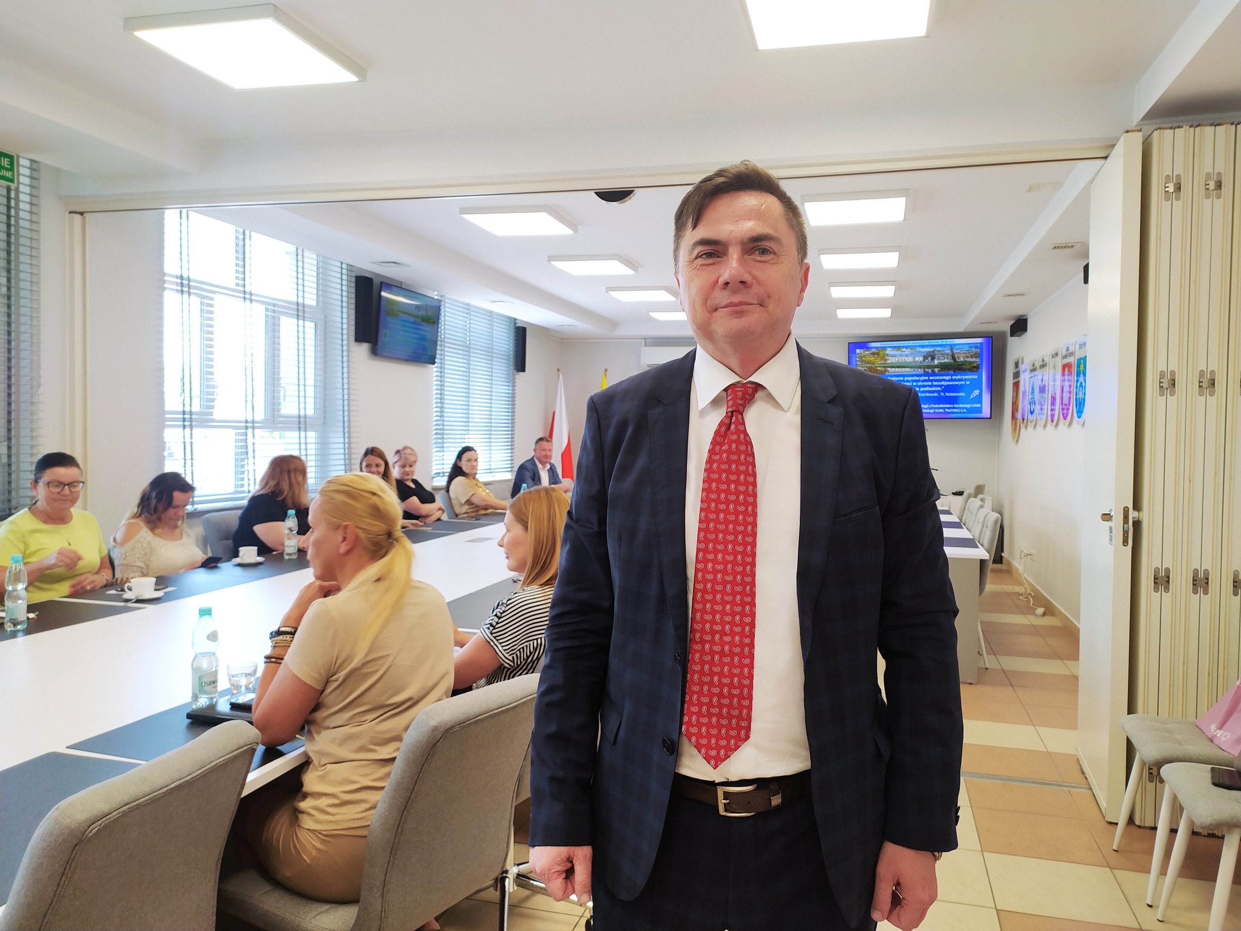 Mężczyzna stoi w sali, w której przy stole siedzą ludzie.