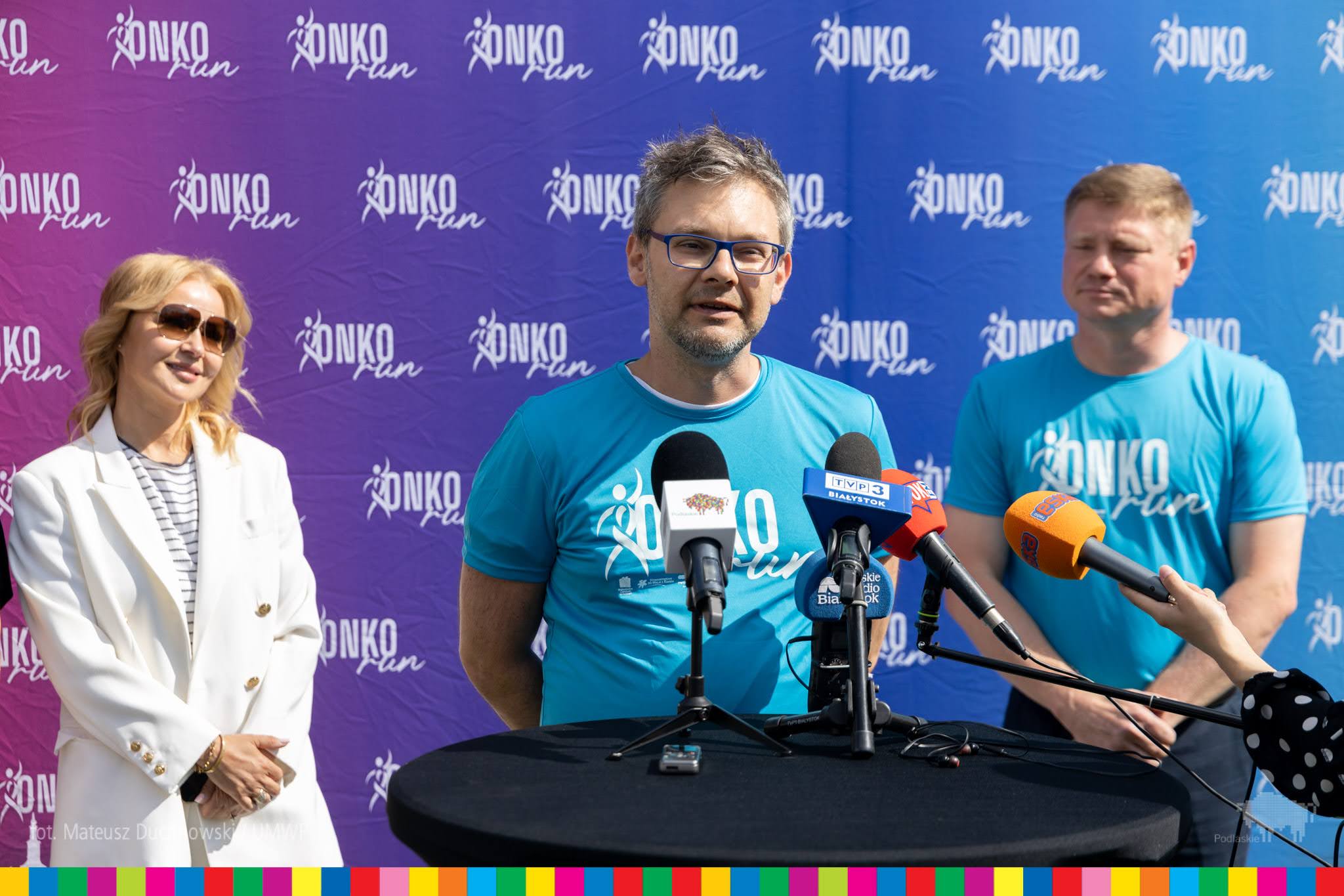 Kobieta w białej marynarce i dwóch mężczyzn w niebieskich koszulkach z napisem onkorun stoją przed mikrofonami