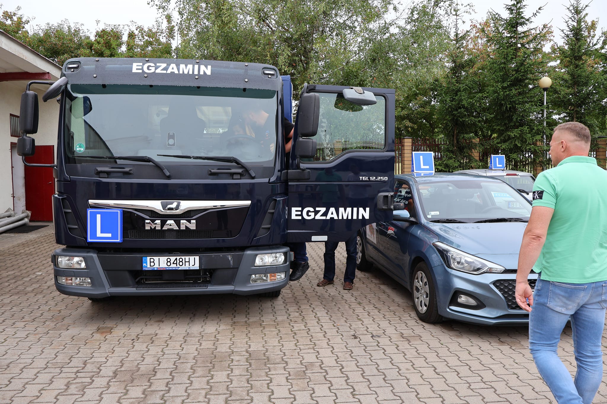 Ciężarówka stoi na placu egzaminacyjnym, obok samochód osobowy, obok przechodzi mężczyzna